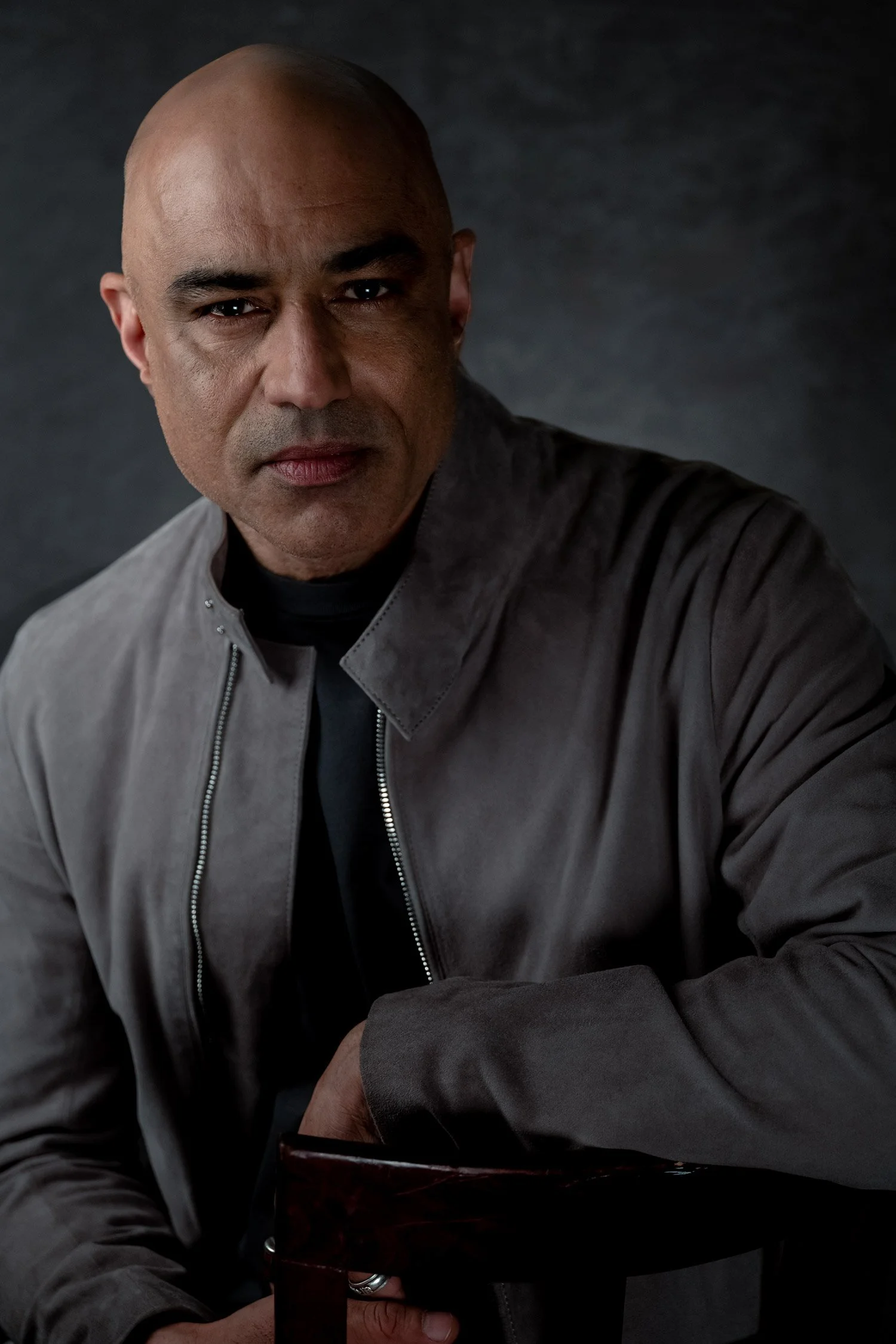 Portrait of theater actor Faran Tahir, seated with arm resting on chair back. He wears a gray suede jacket over a black sweater, looking intently into the camera while bathed in dramatic, contrasty light against a bluish, dark, mottled backdrop.
