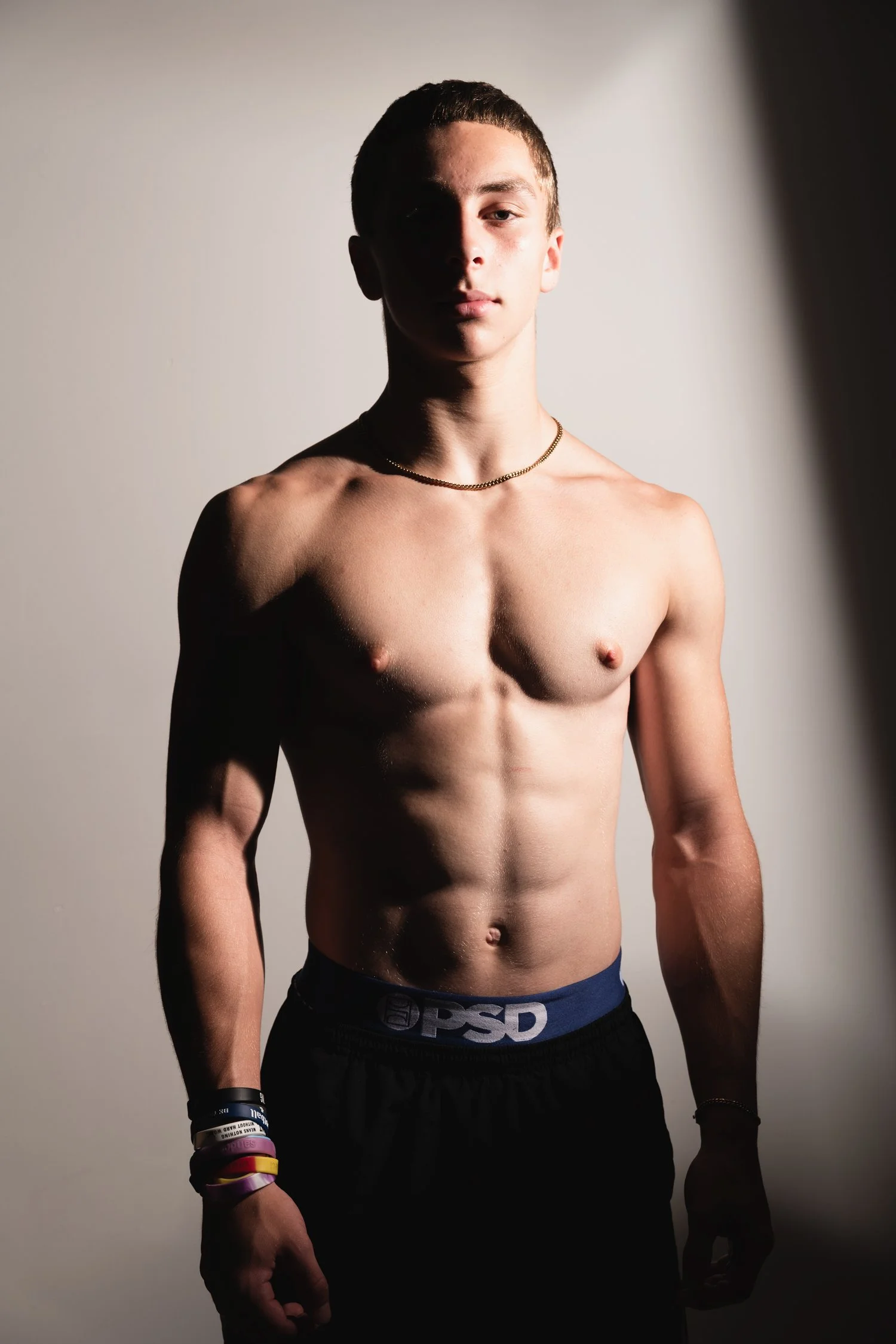 A shirtless young man standing with an intense expression, wearing black shorts, a gold chain necklace, and multiple colorful wristbands, with dramatic lighting casting one side of his face and body in shadow.
