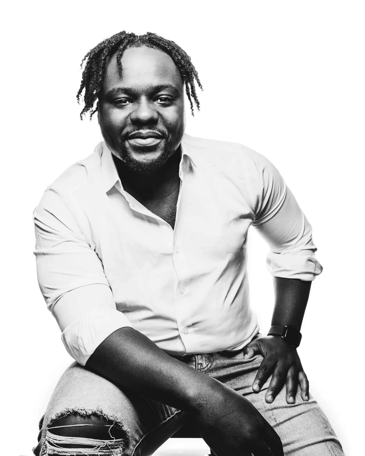 Young Black man in dreads, white shirt and torn jeans, seated on a stool, leaning in toward the view. Portrait is in black and white against a white backdrop.