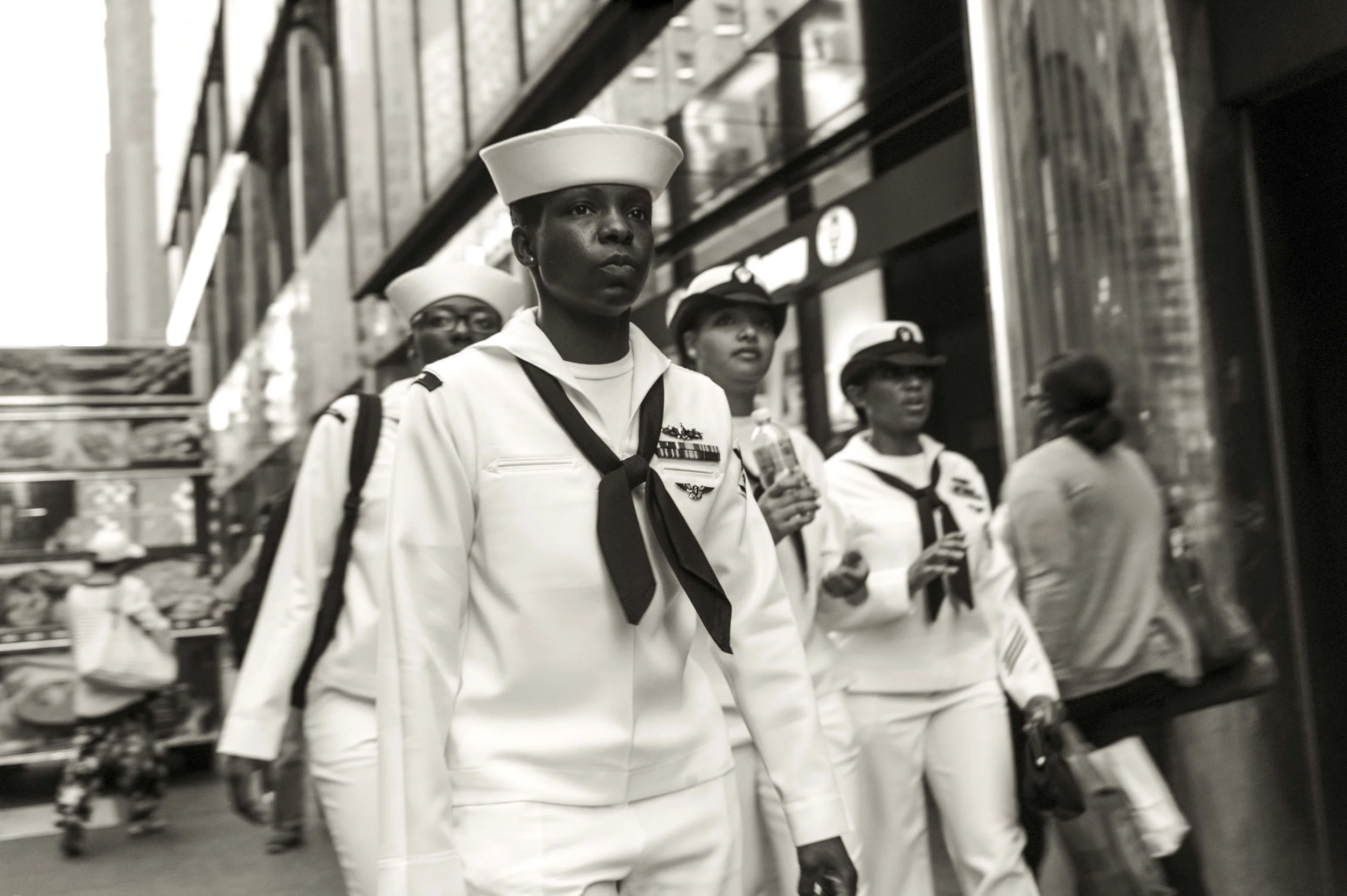 A group of young women in U.S. Navy uniforms participating in a parade or march, walking along a street lined with shops and street vendors.