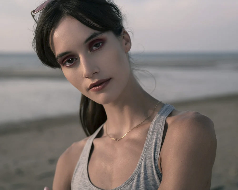 A young woman with dark hair and makeup, wearing a gray tank top and a delicate gold necklace, standing on a beach with waves and a cloudy sky in the background.