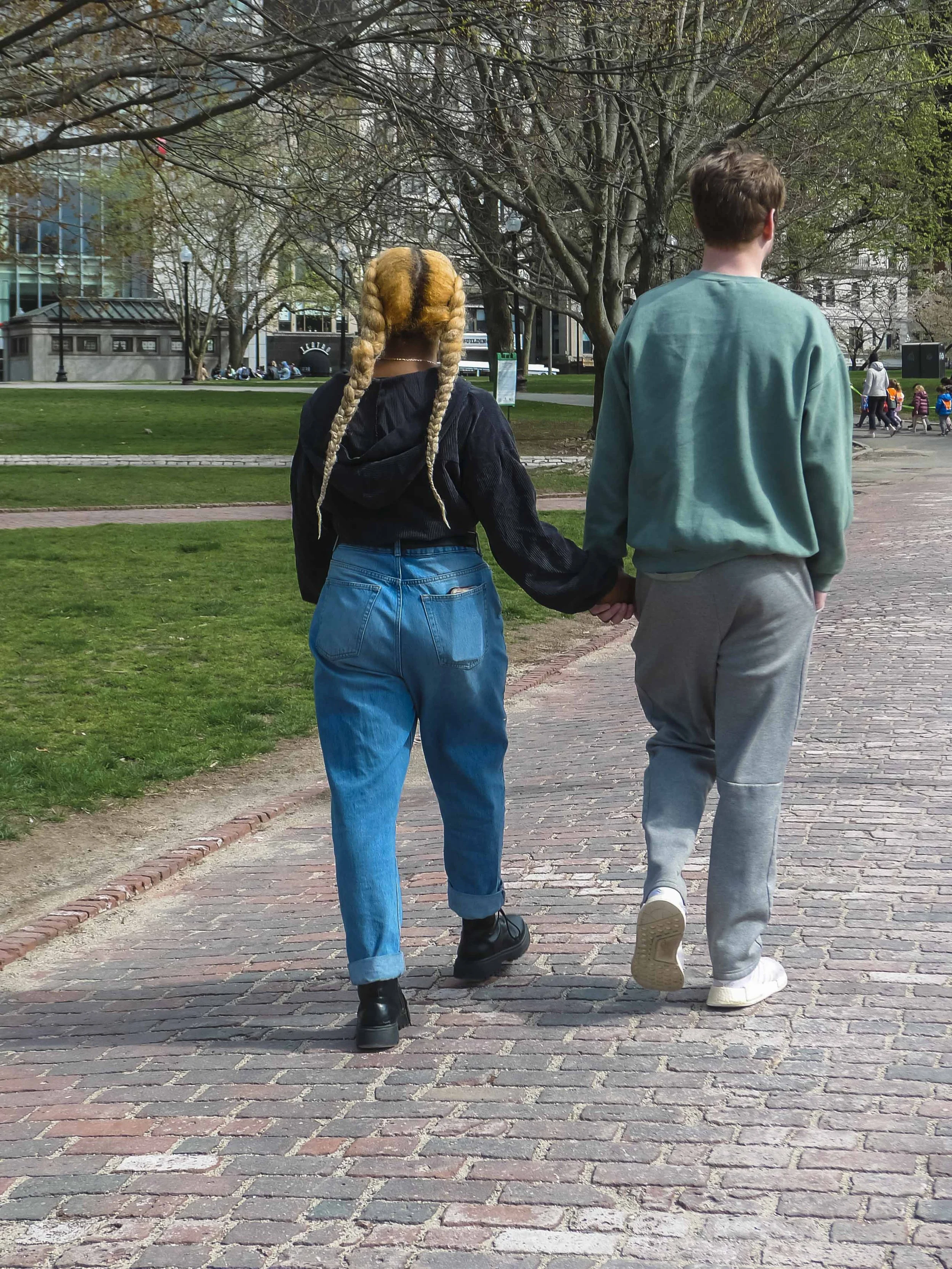 A young woman and a young man walking hand in hand through a park, with trees and buildings in the background.