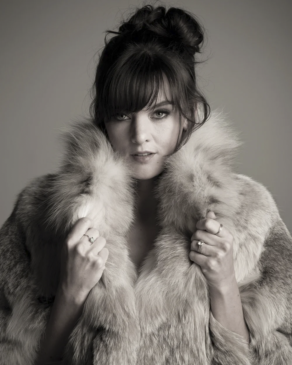 Woman with dark hair in an updo, wearing a fur coat, looking at the camera.