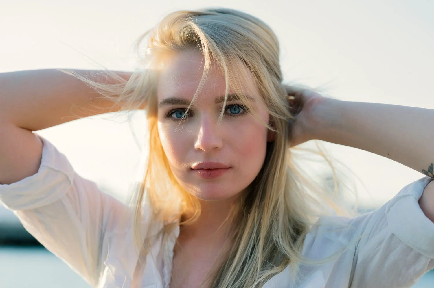 A young woman with blonde hair and blue eyes looking directly at the camera, with her arms behind her head, outdoors during golden hour.