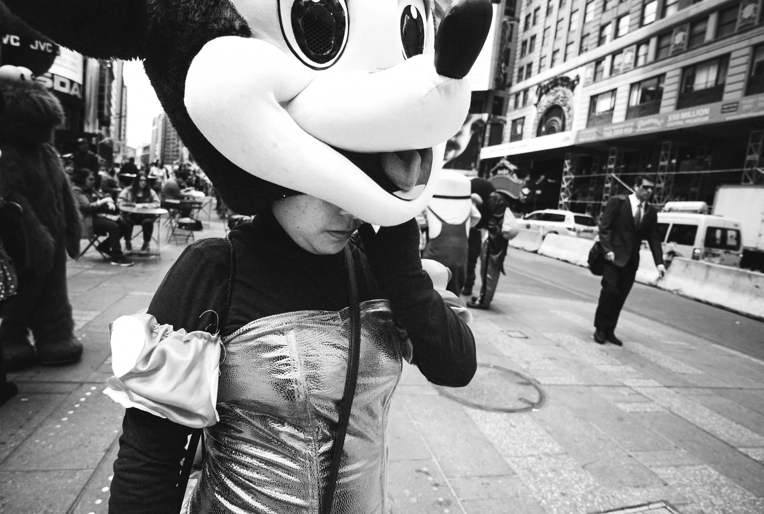 Person wearing a large cartoon character mask with big eyes and tongue sticking out, walking on a city sidewalk with buildings and other pedestrians in the background.