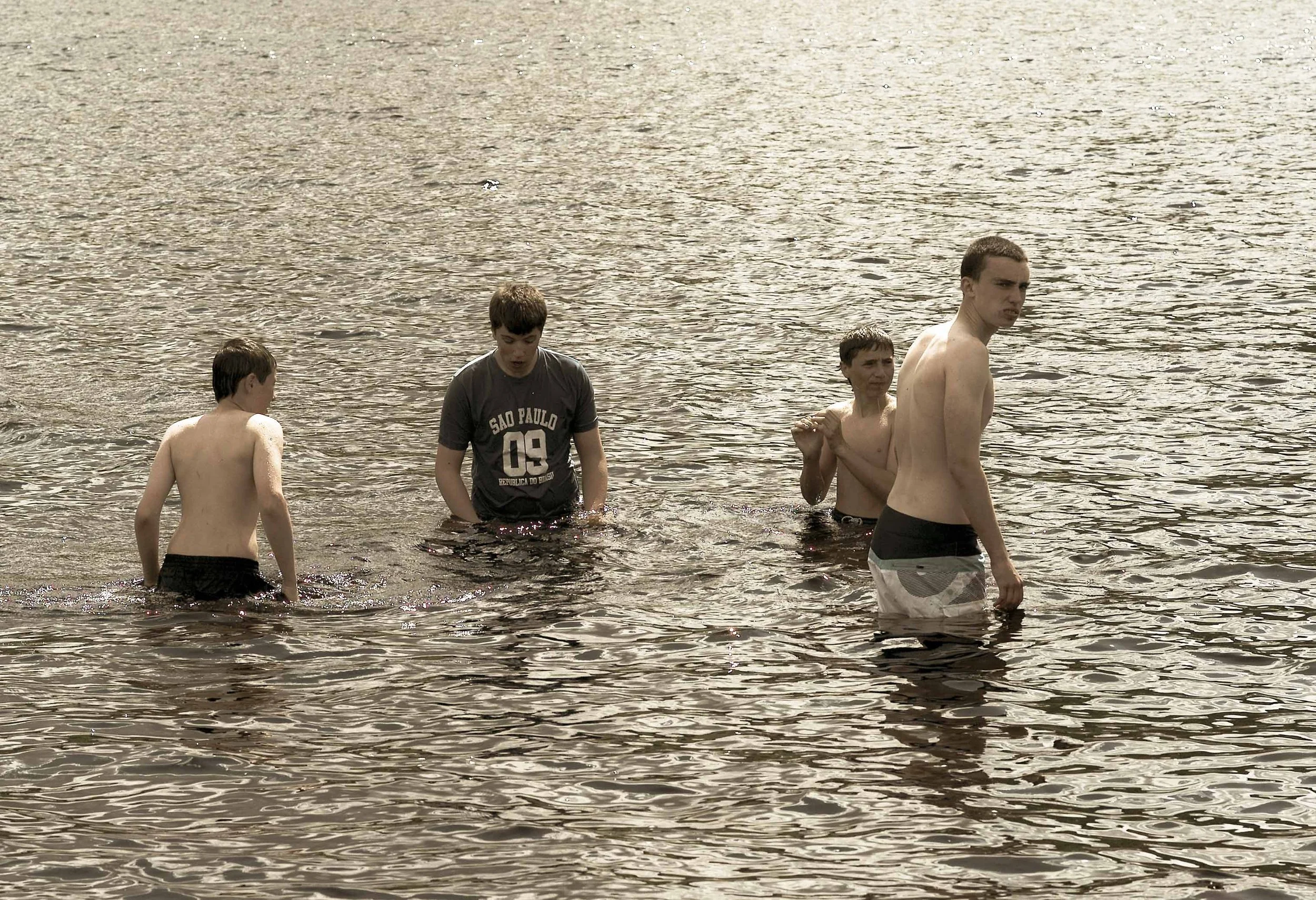 Four young males standing in water at a beach or lake, one shirtless, one wearing a dark T-shirt, and two others shirtless, with the water reaching their waists.