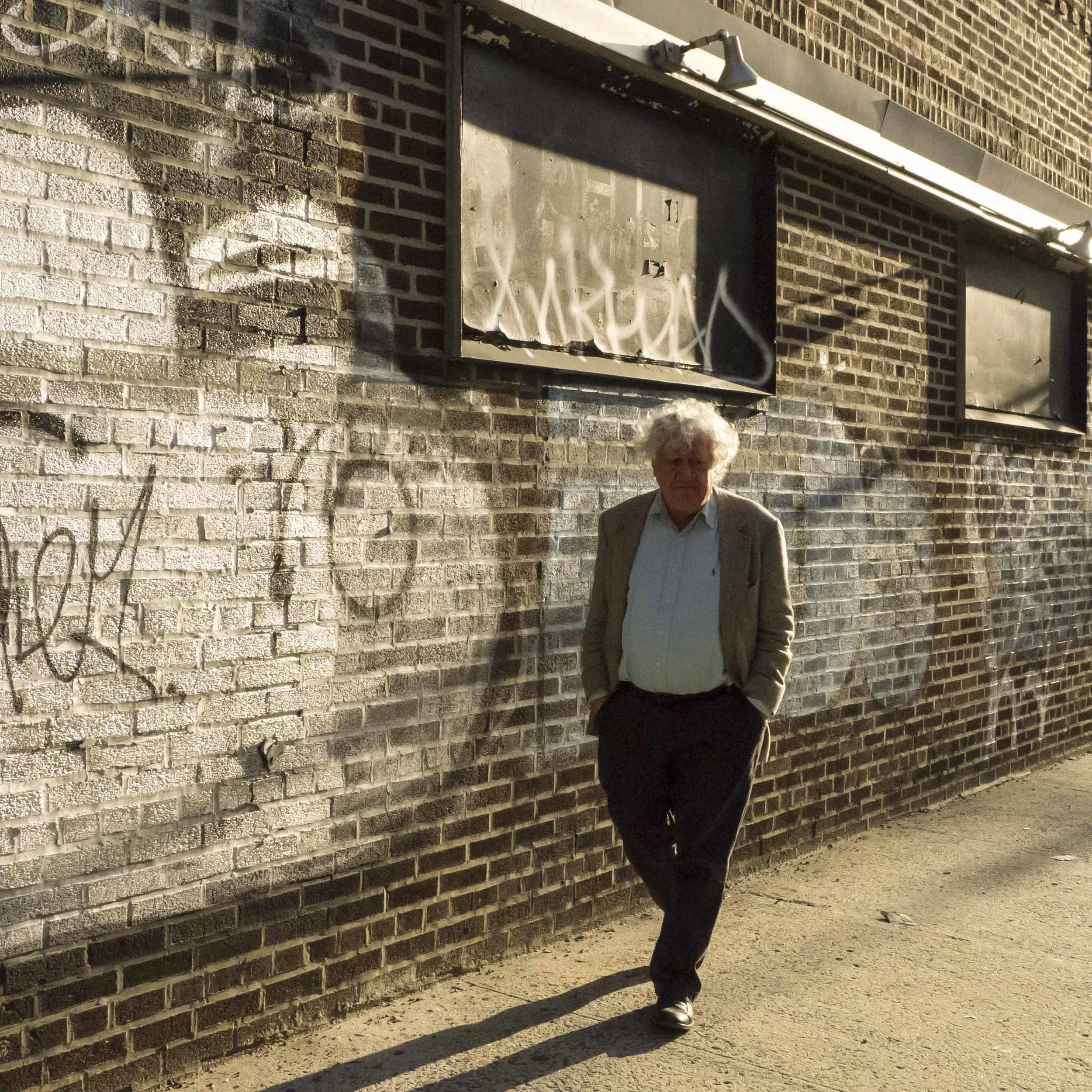 A man with white curly hair wearing a brown blazer and light-colored shirt walking along a brick wall and sidewalk, with graffiti and shadows visible on the wall.