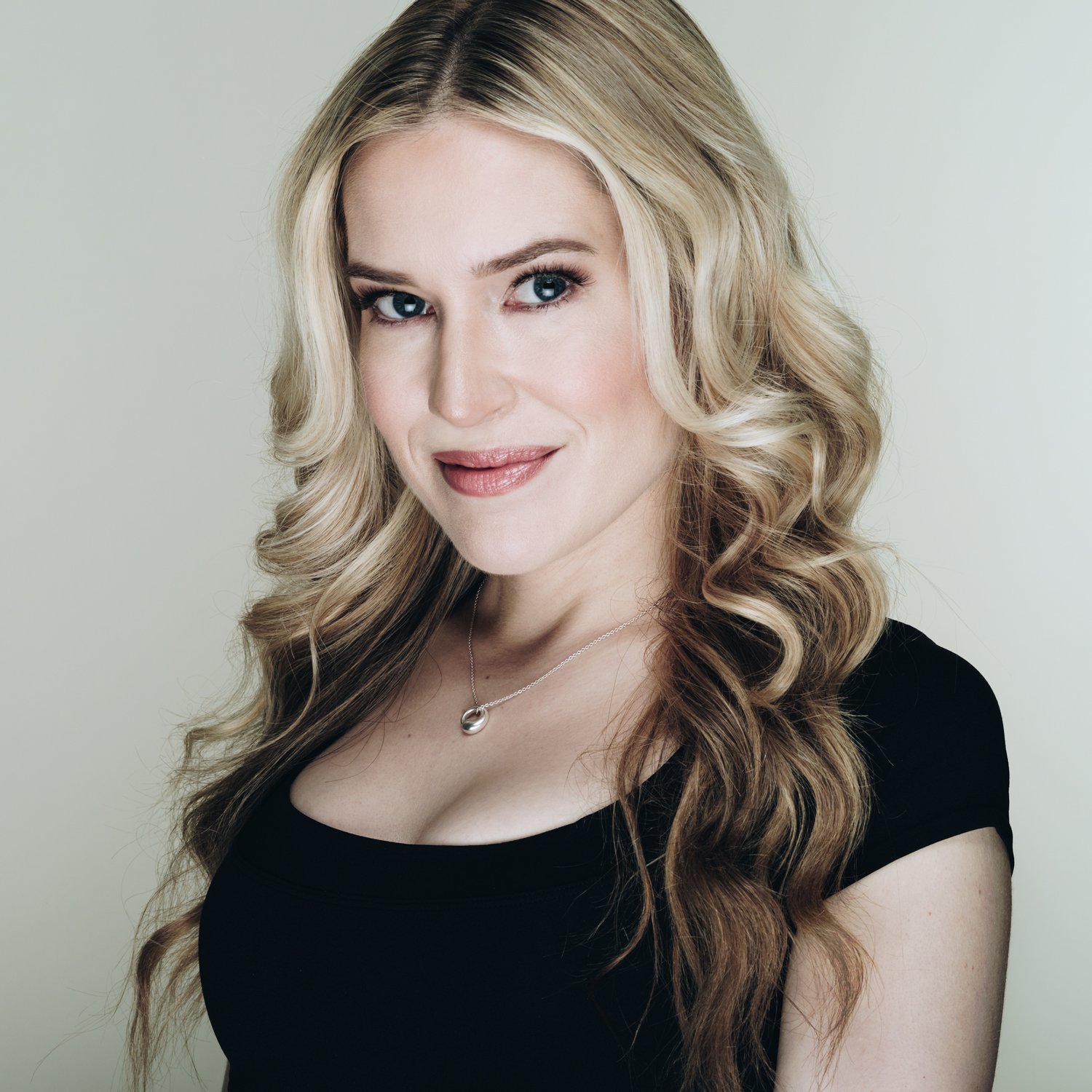 Professional headshot of a young woman with long, blonde hair wearing a black, low-neck, cap-sleeve top against a light blue-gray background.