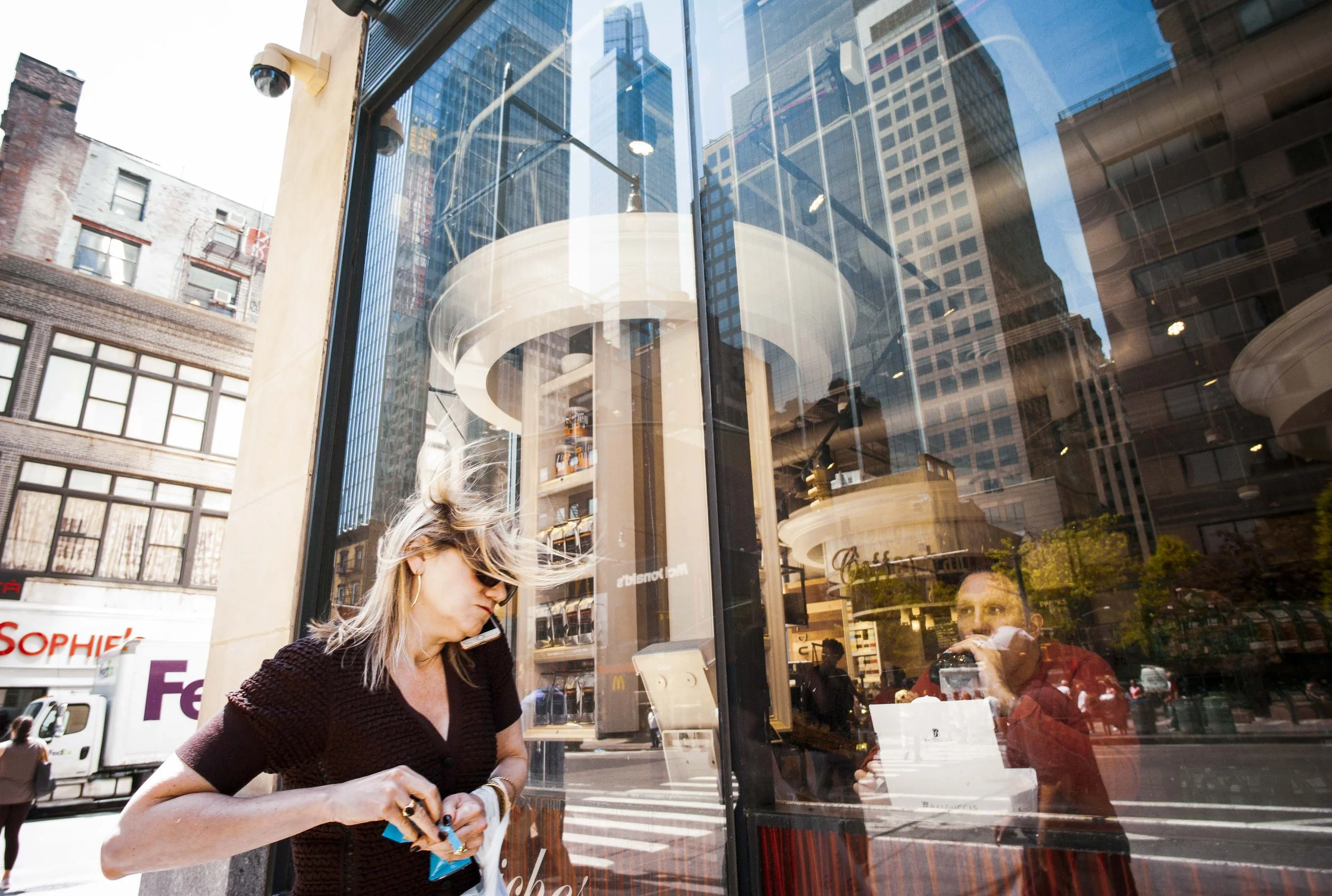 A woman with blonde hair and sunglasses is standing outside a store, looking at her phone. The store has a large glass window reflecting surrounding buildings and cityscape, with several people visible inside and outside.