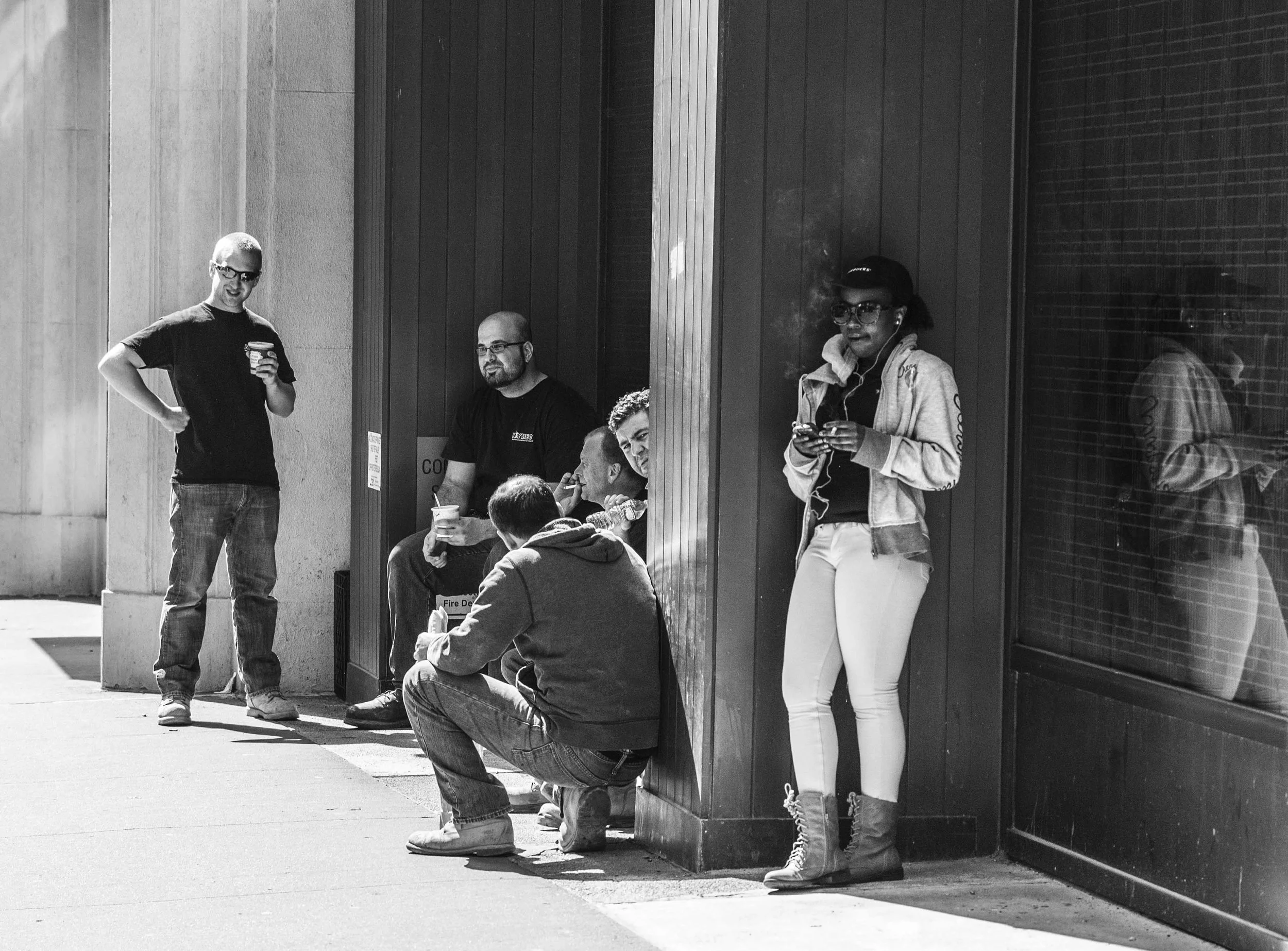 A group of four people, three men and one woman, standing or sitting outside a building. The woman is wearing sunglasses and earphones, holding a smartphone, while one man is sitting on the ground with a beverage, and two men are standing nearby, one
