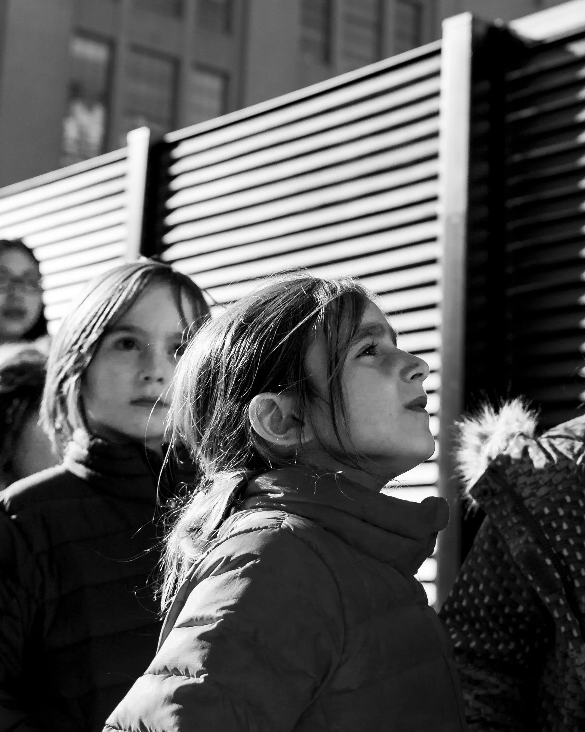 Black and white image of two women with long hair looking upward in an outdoor urban setting with a building and striped panel in background.