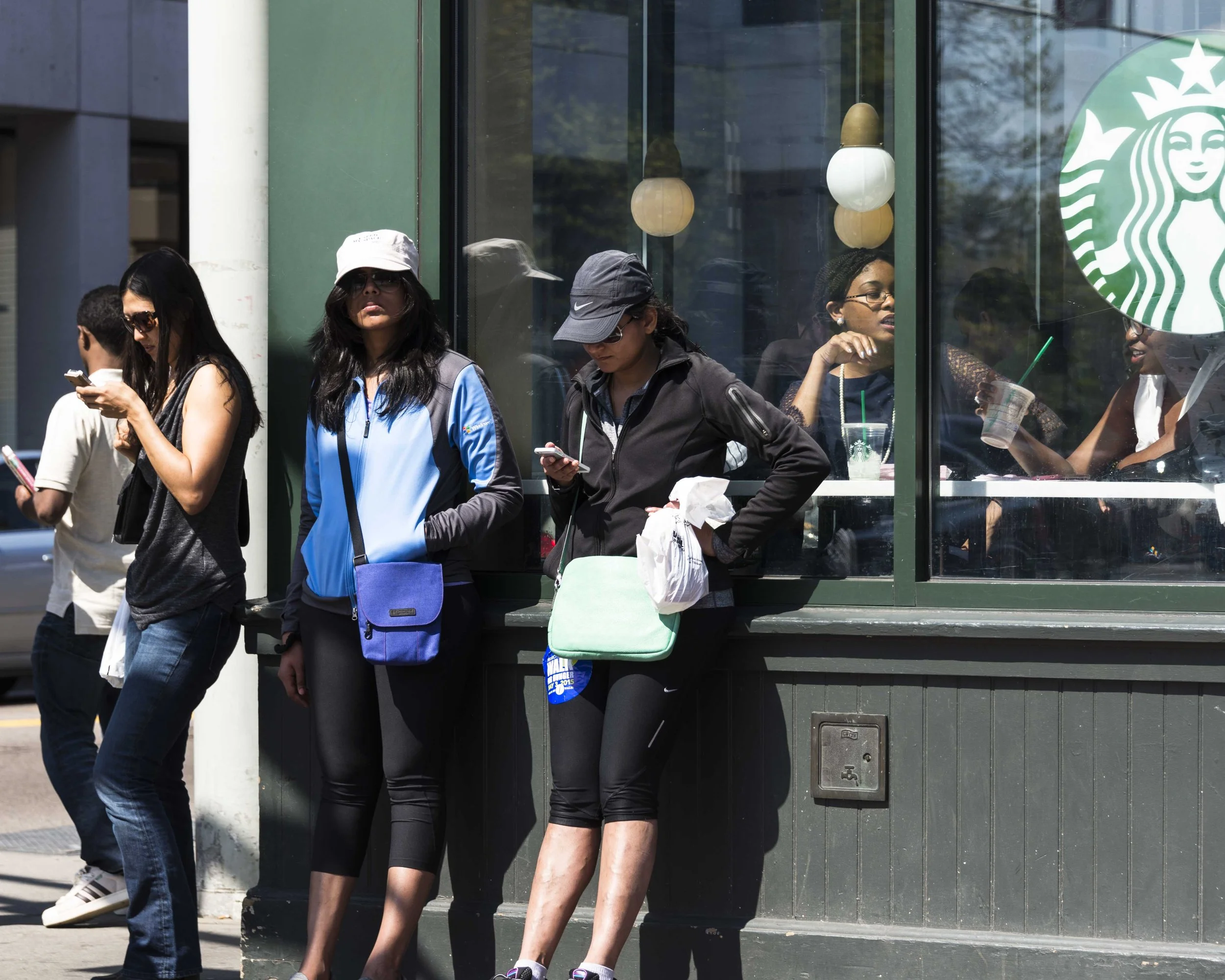 Four women waiting outside a Starbucks coffee shop, some looking at their phones, one holding a coffee cup, under hanging lights, with a Starbucks logo window.