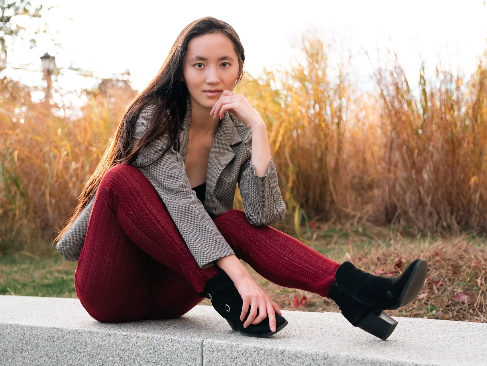 A young woman with long dark hair, sitting on a concrete ledge outdoors during fall, wearing a gray blazer, maroon pants, and black boots.