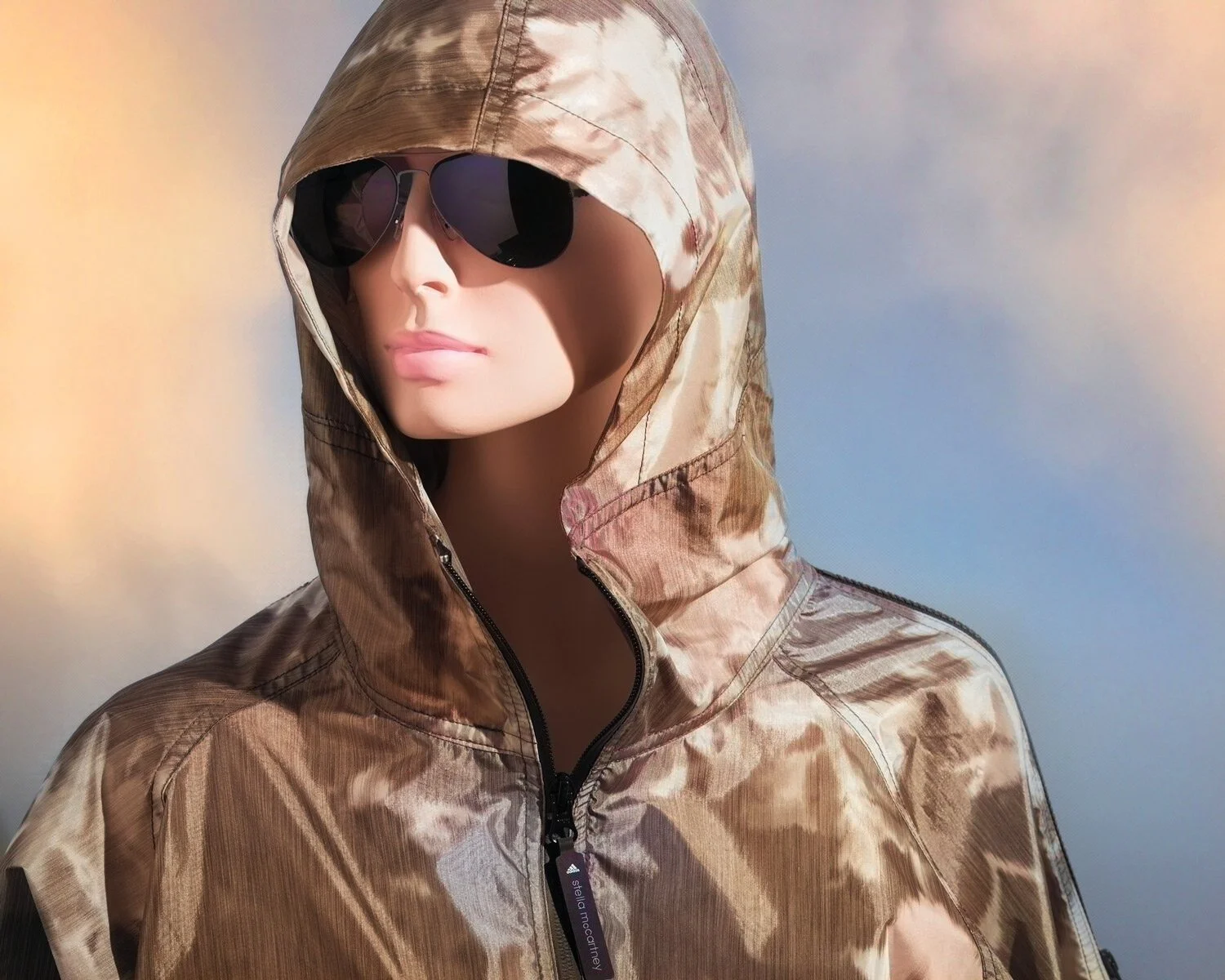 A mannequin wearing a camouflage-patterned hooded jacket and black sunglasses against a cloudy sky background.