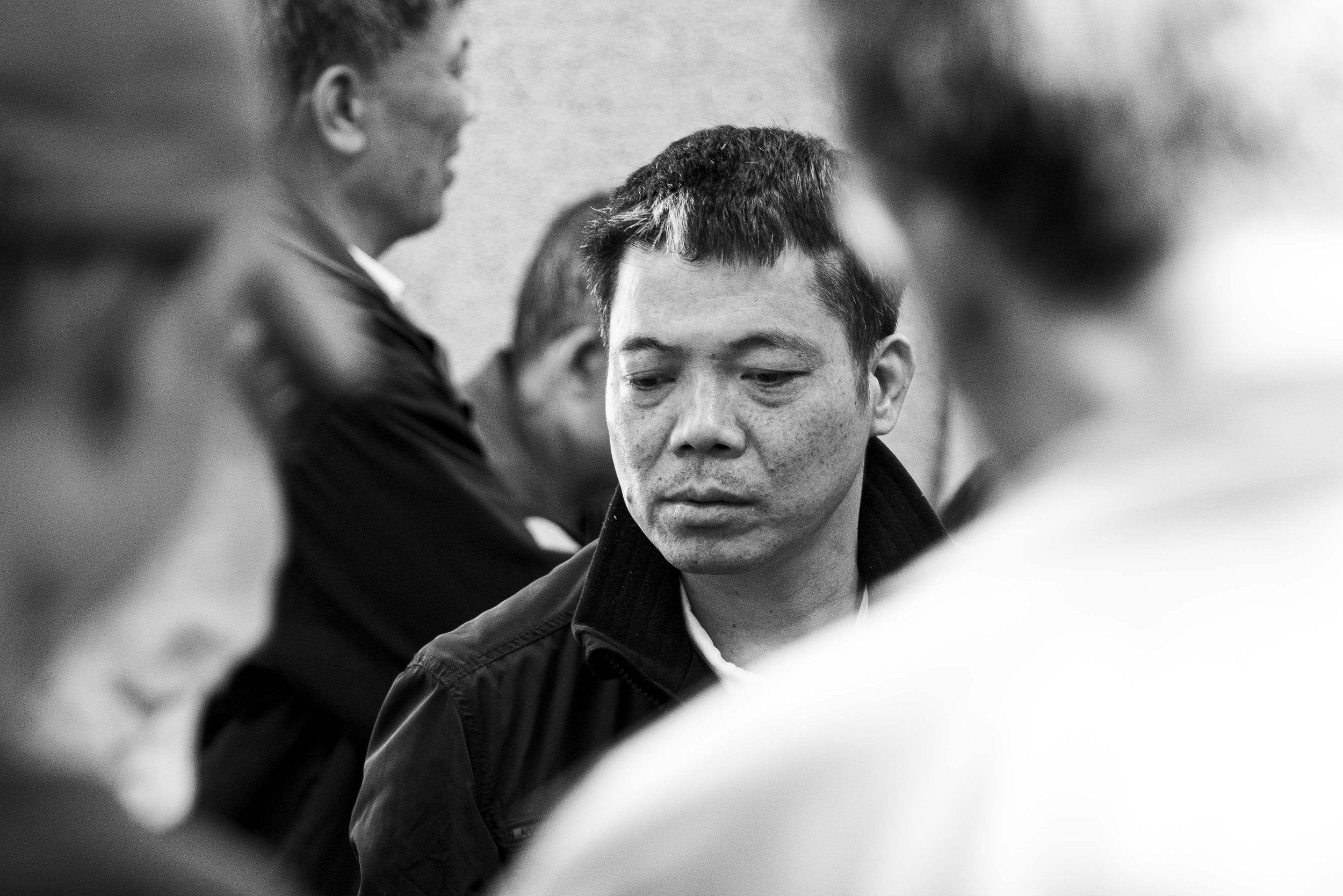 Black and white photo of a young man with short hair looking down, surrounded by blurred people in a group setting.