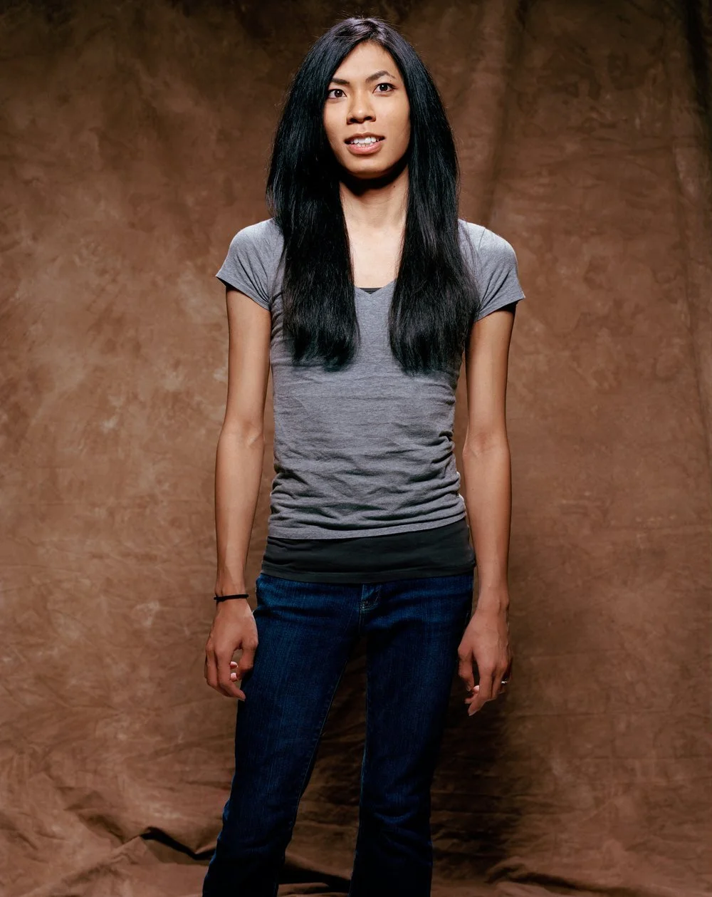 Young woman with long black hair, wearing a gray t-shirt and jeans, standing against a brown textured background.