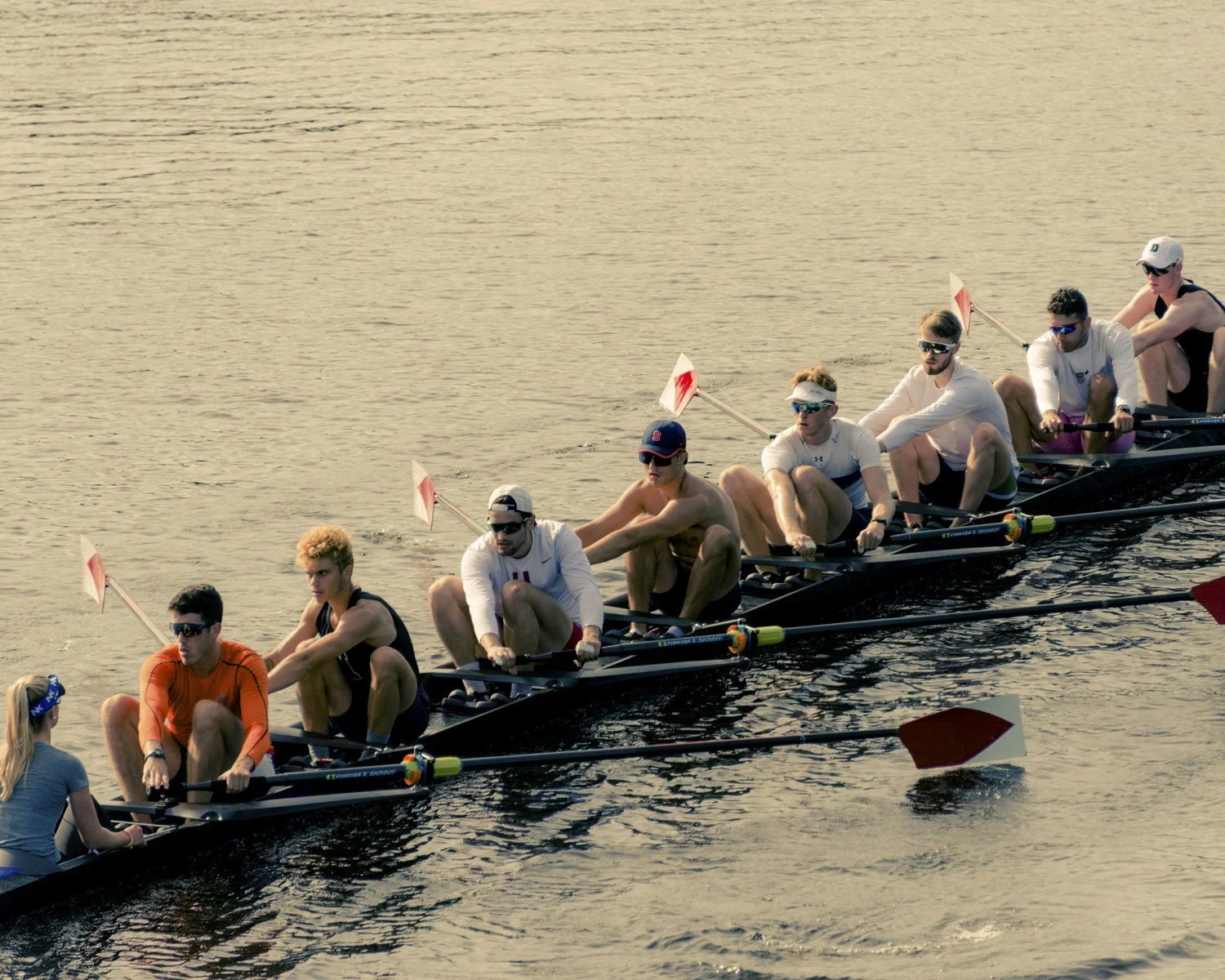 A group of male and female rowers in a racing shell, competing in a rowing race on a body of water during sunset or late afternoon.