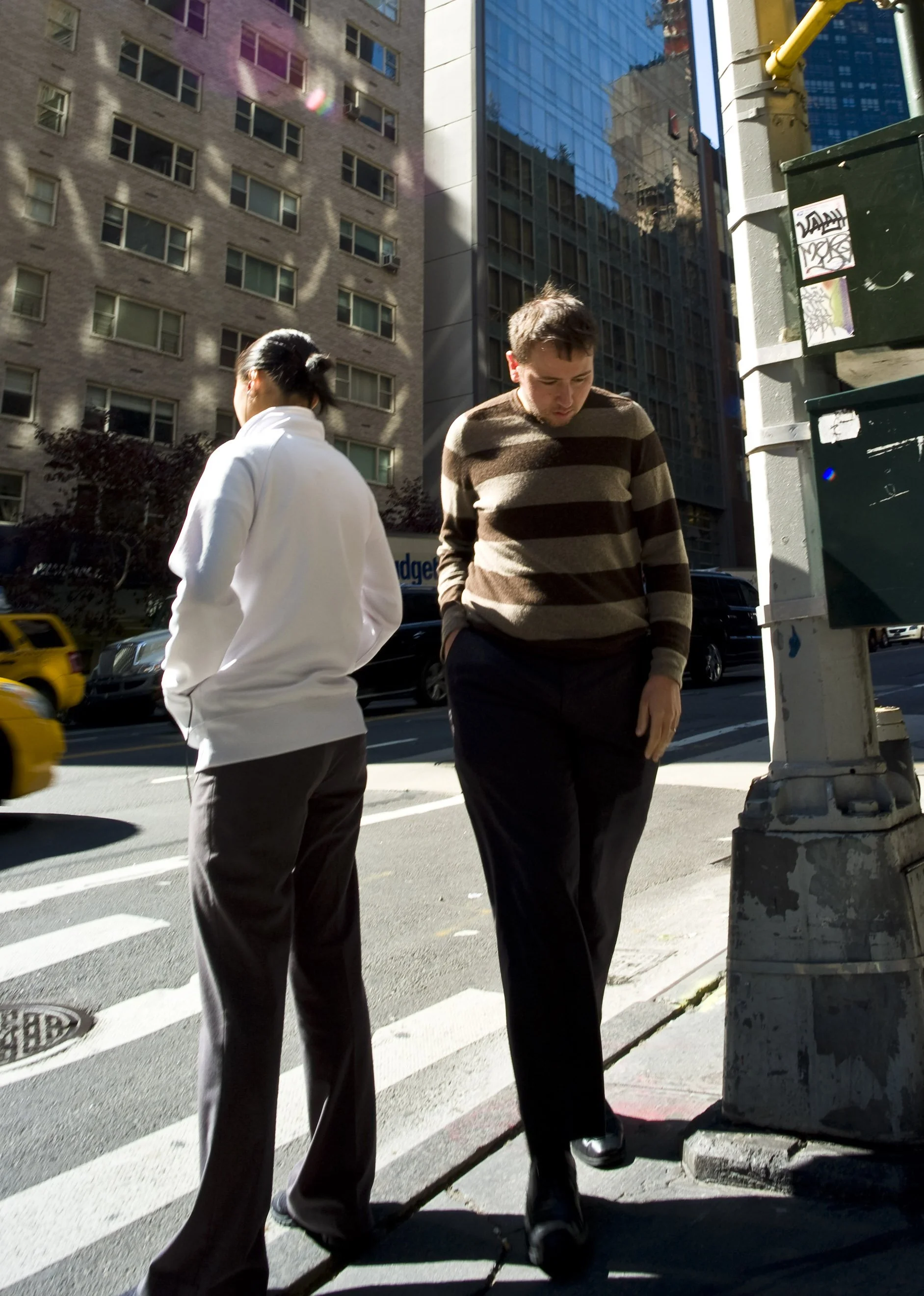 Two men standing on a city sidewalk near a crosswalk, with tall buildings and a reflective glass building in the background, one with his hands in his pockets and the other with his head bowed.