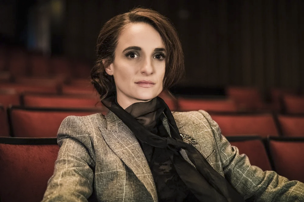 A woman with short brown hair sitting in an empty theater with red seats, wearing a beige checkered blazer and a black scarf, looking directly at the camera.