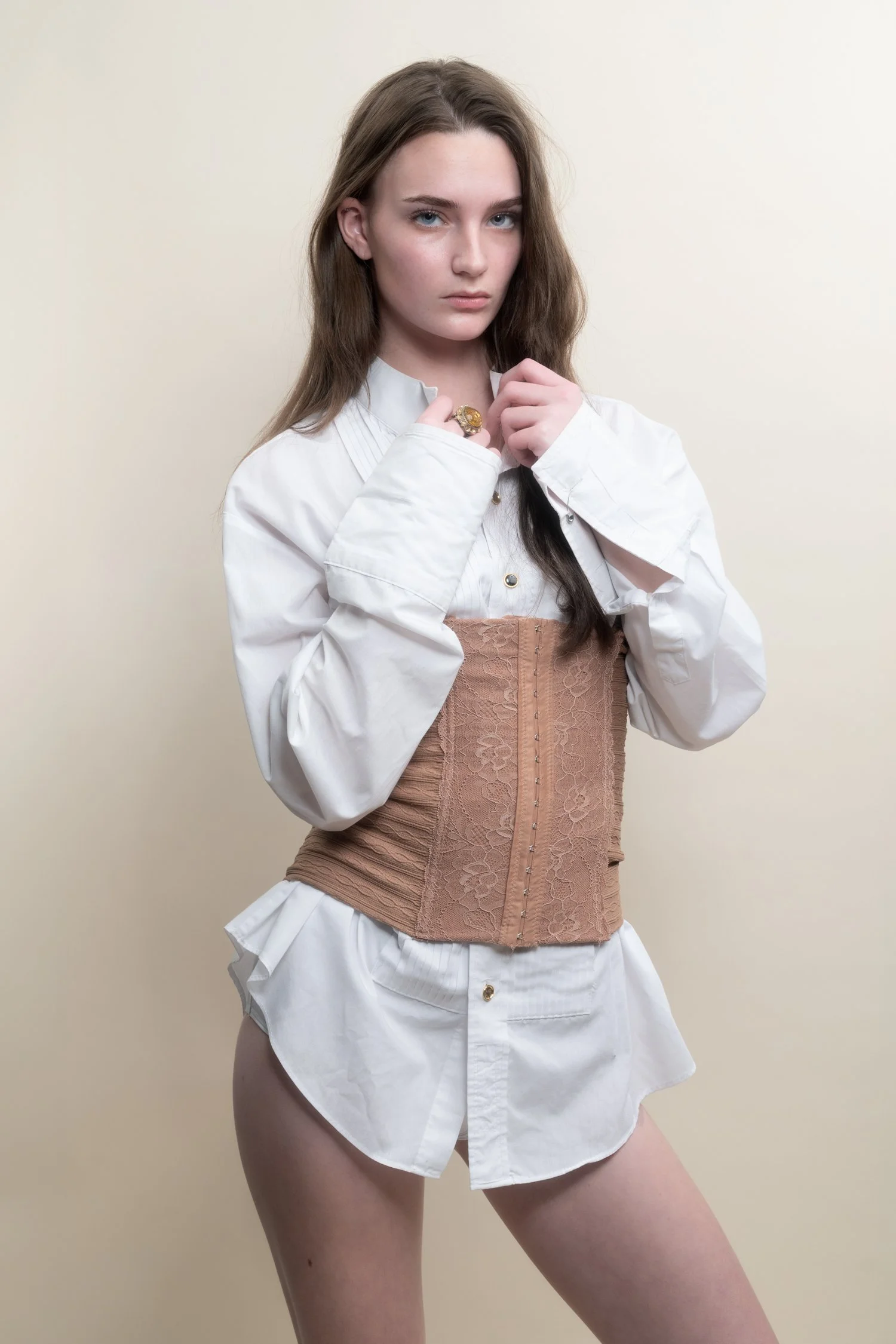 Young woman with long brown hair wearing a white shirt, a tan lace corset, and white shorts against a plain beige background.