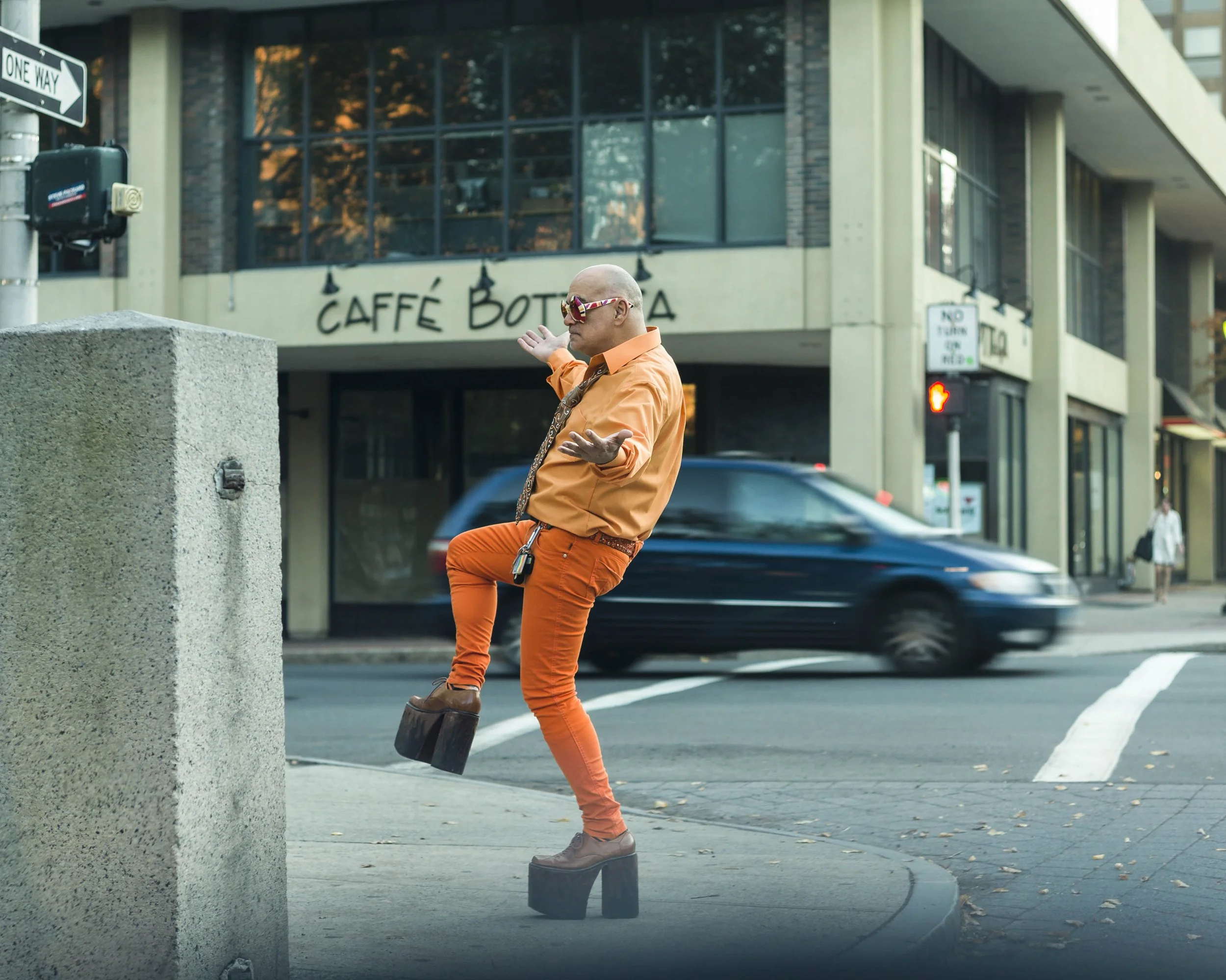 A man with a bald head and sunglasses wearing an orange shirt and pants poses on a city sidewalk, with his arms open and one leg raised, wearing high-heeled platform shoes from the 1970s.