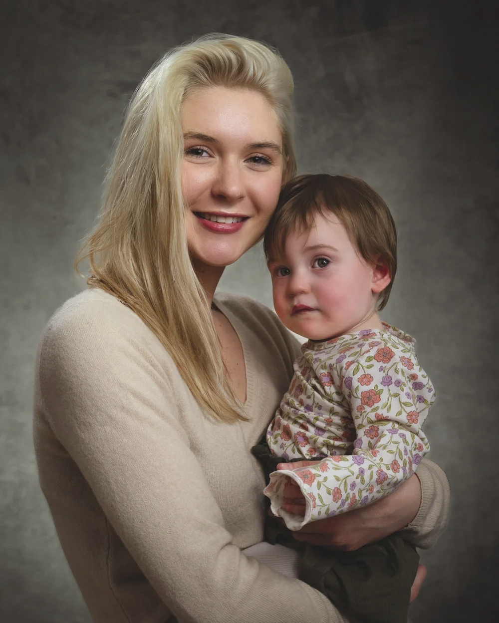 A woman with blonde hair smiling and holding a young child with brown hair against a gray background.