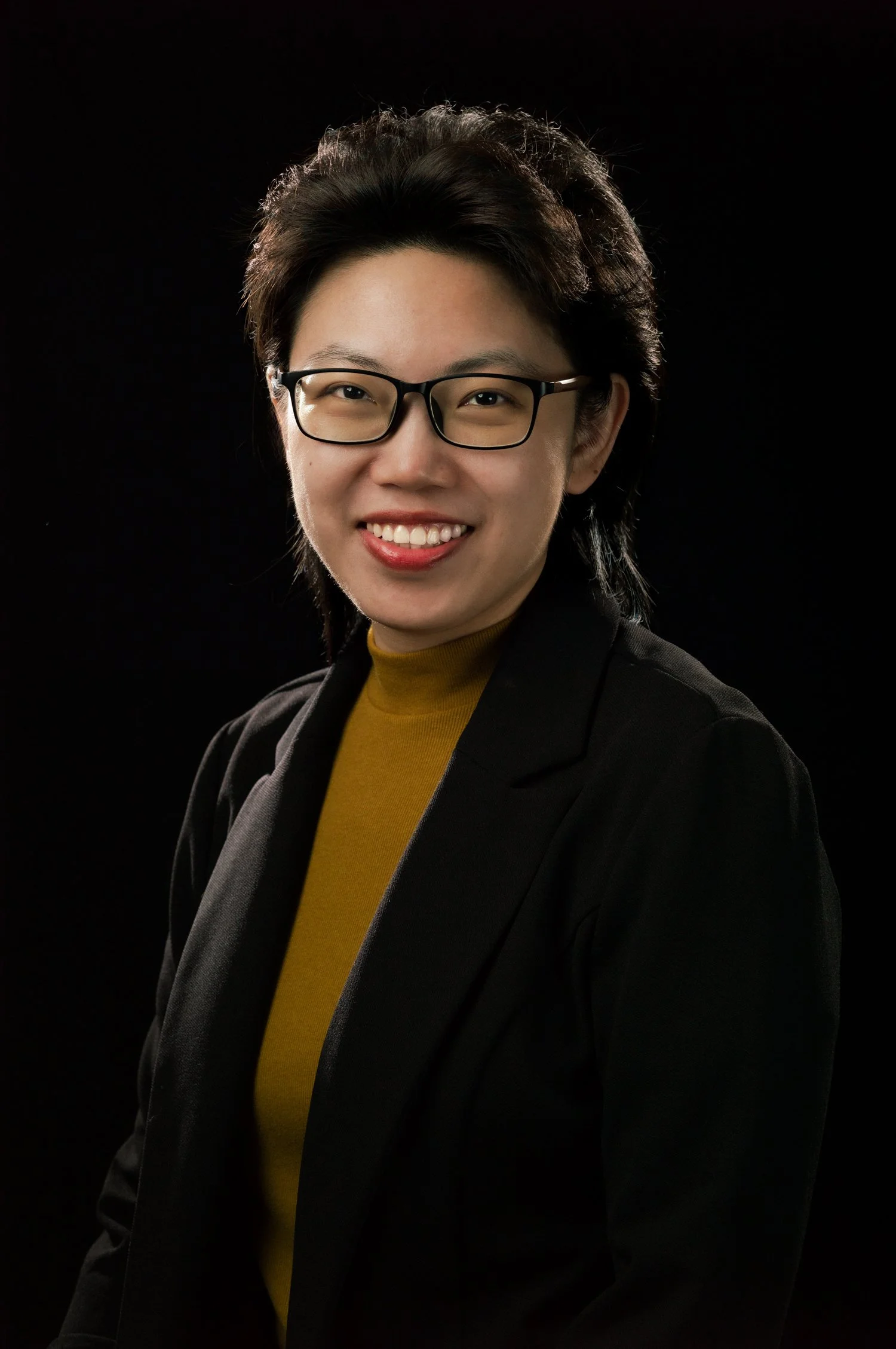 Professional portrait of a woman with glasses, wearing a black blazer and gold turtleneck, against a dark background.
