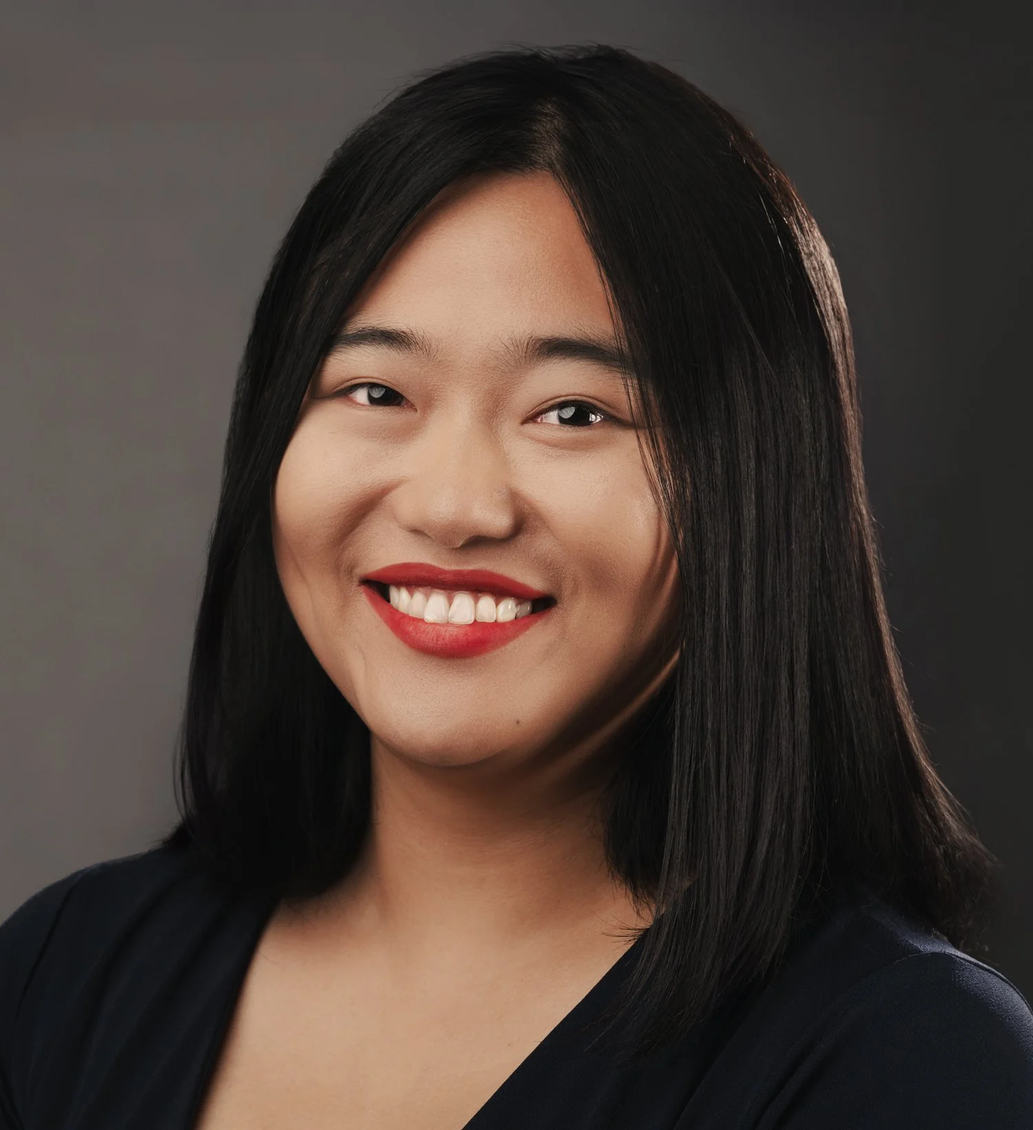 Studio headshot of smiling young Asian woman with shoulder-length straight, black hair photographed on a simple gray background.