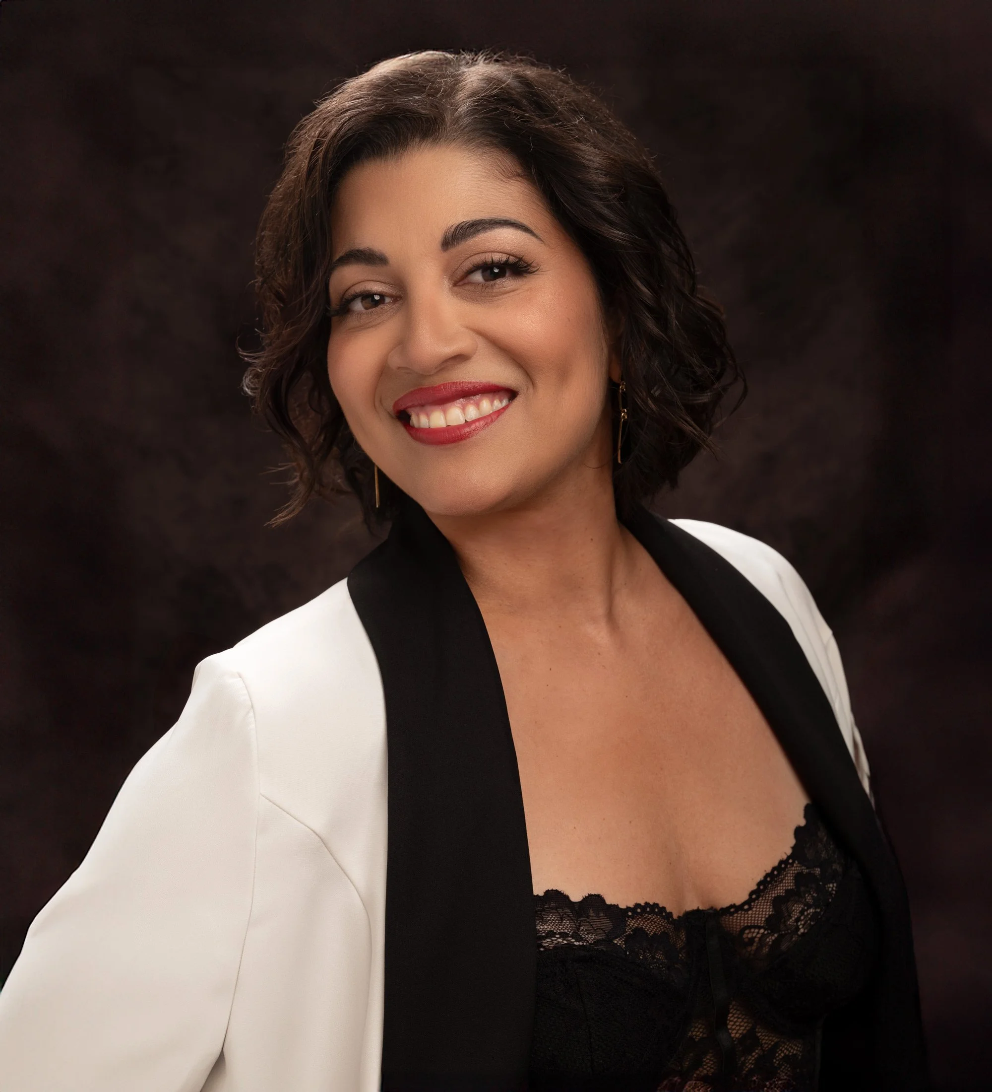 Portrait of smiling, expressive Latina woman with dark hair styled in a bob. She has bright red lips and wears a white and black jacket over a black lace lingerie top. The background is dark brown oil cloth and she is cropped just below the breast.