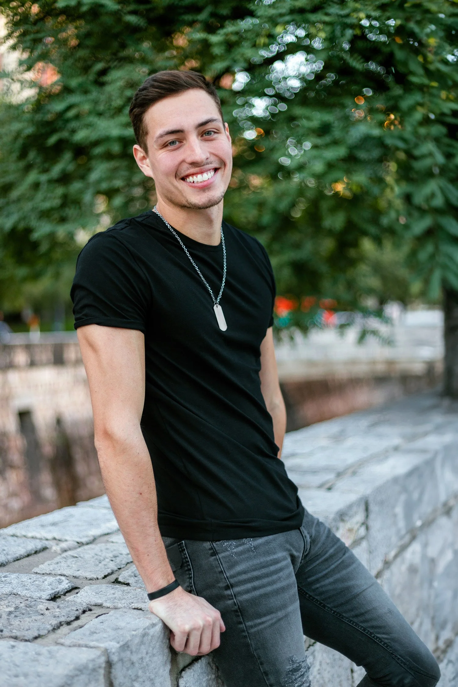 Smiling young man in a black t-shirt and blue jeans, leans against a short, stone wall with trees in background.