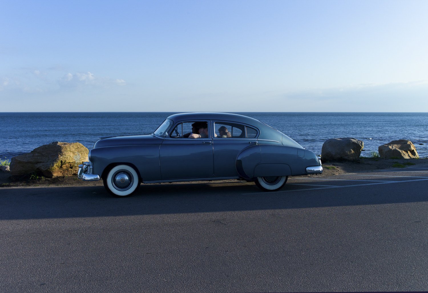 A vintage blue car parked beside rocks overlooking the ocean on a clear day.