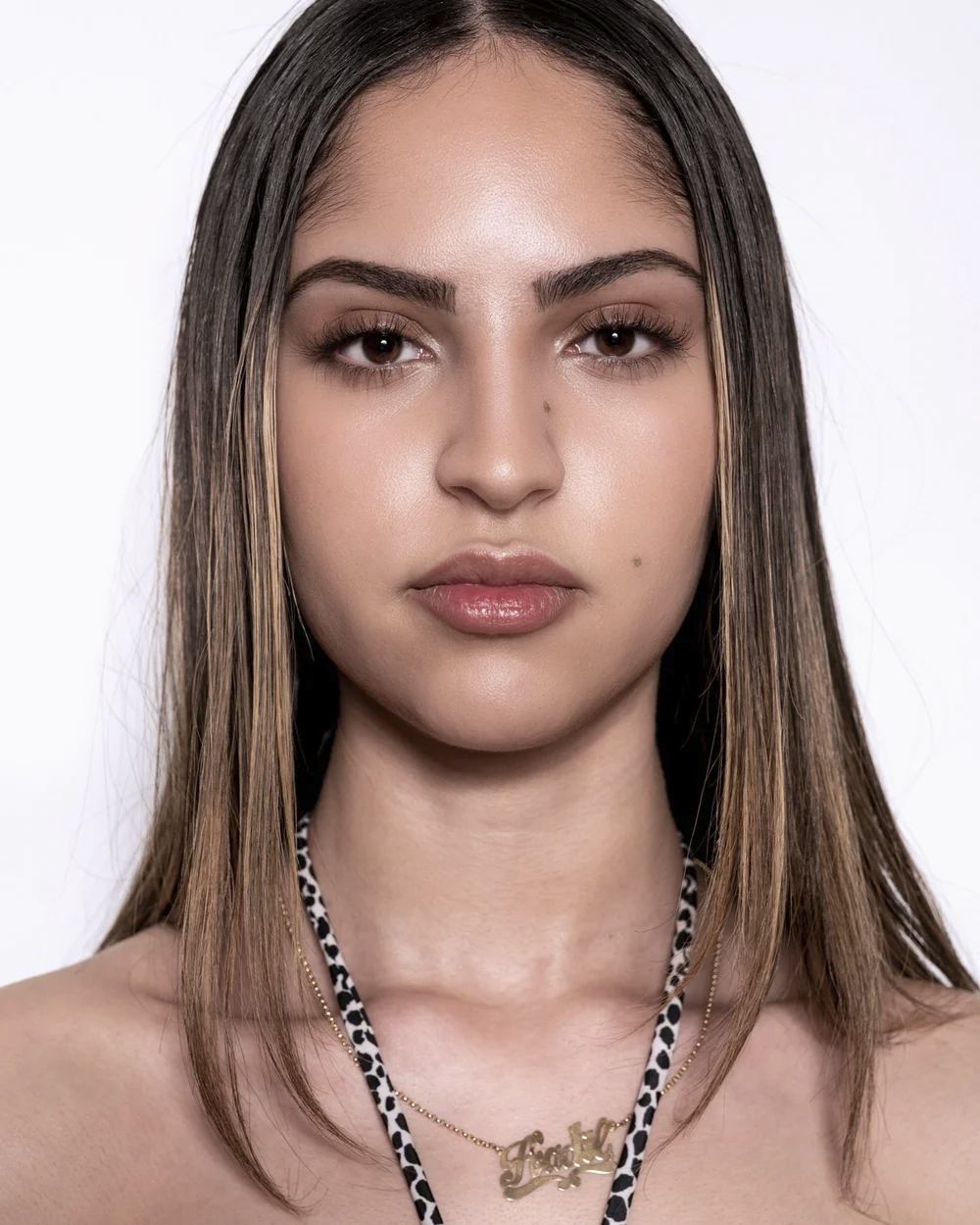 Close-up of a young woman with long brown hair, wearing natural makeup and a necklace that spells 'Hadil'. She has a neutral expression and is against a plain white background.