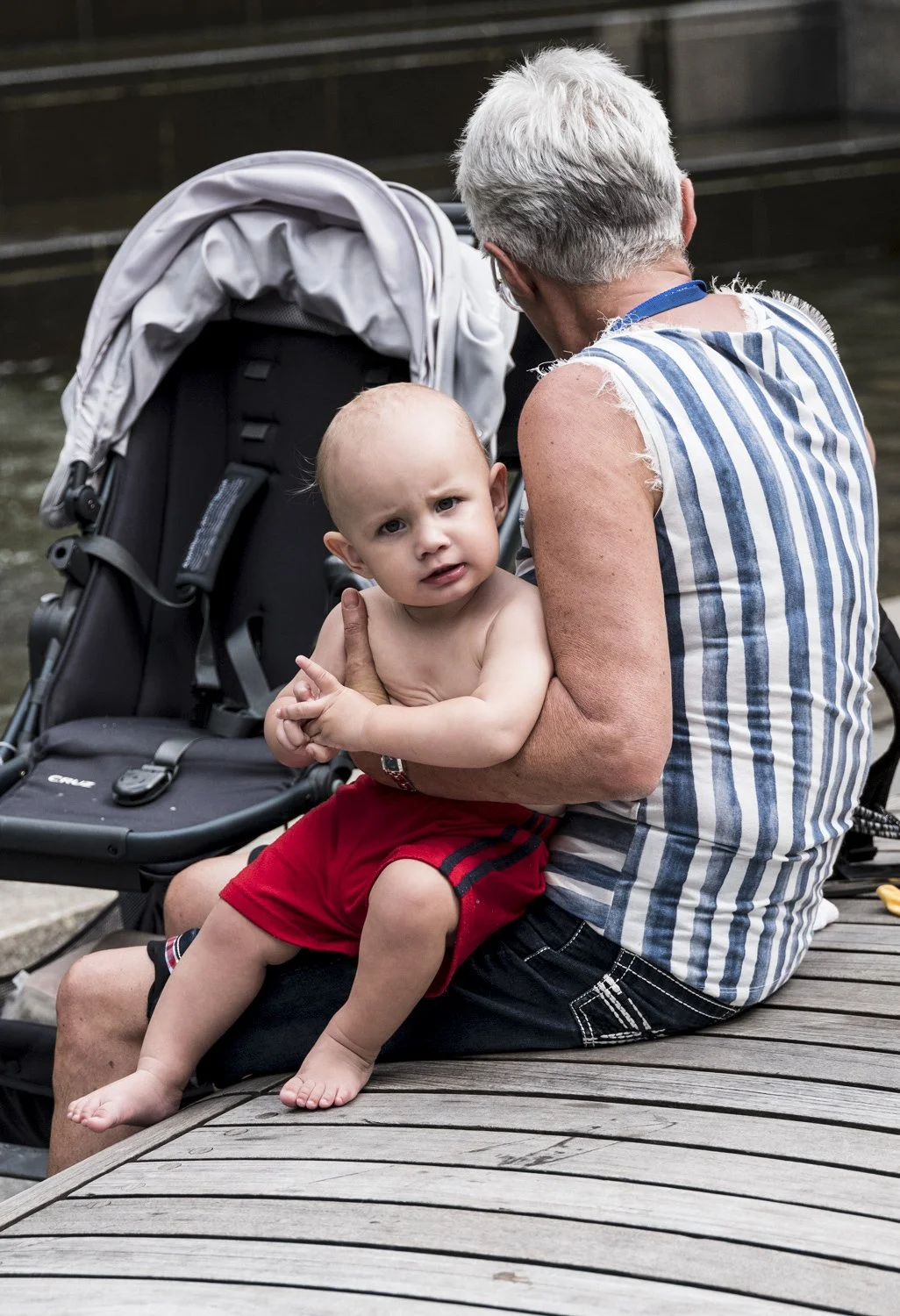 An older woman with short grey hair is sitting on a wooden dock near water, holding a young shirtless boy with red shorts on her lap, the boy looking confused or curious.