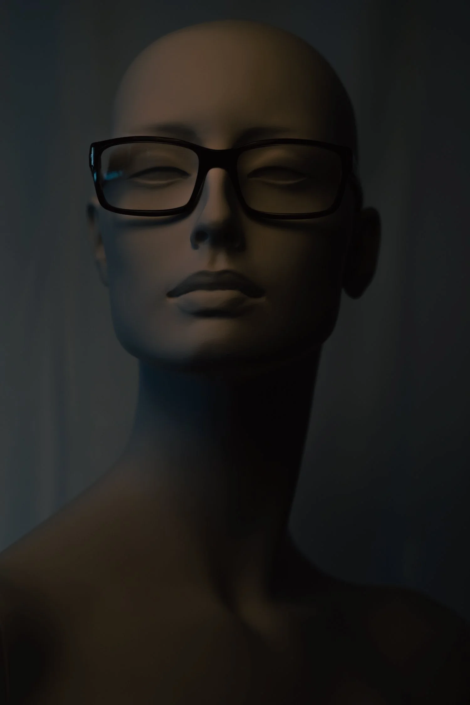 Close-up of a mannequin head wearing black-rimmed glasses, in a dark setting.
