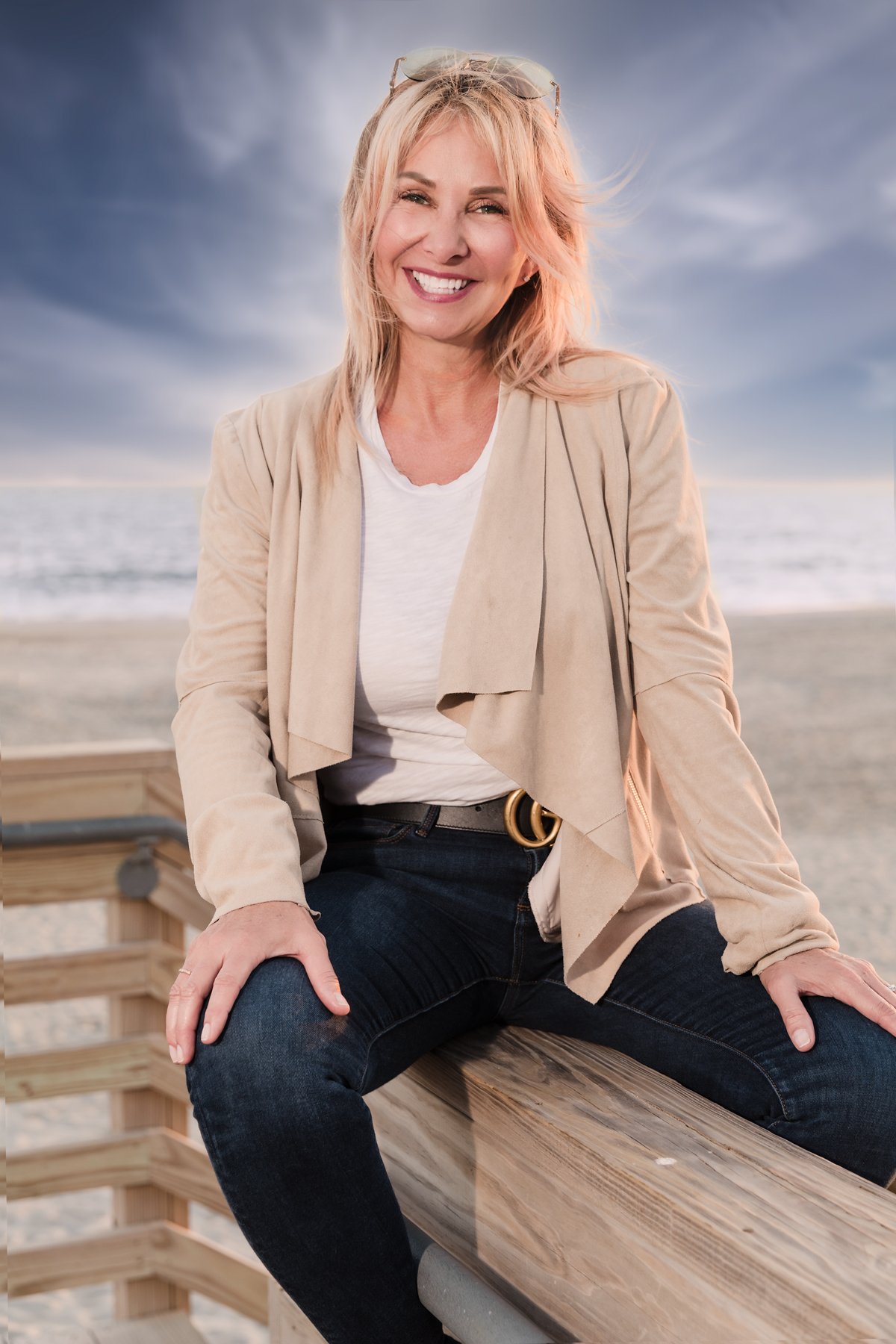 Woman sitting on a wooden bench at the beach, smiling with ocean and sky in the background.