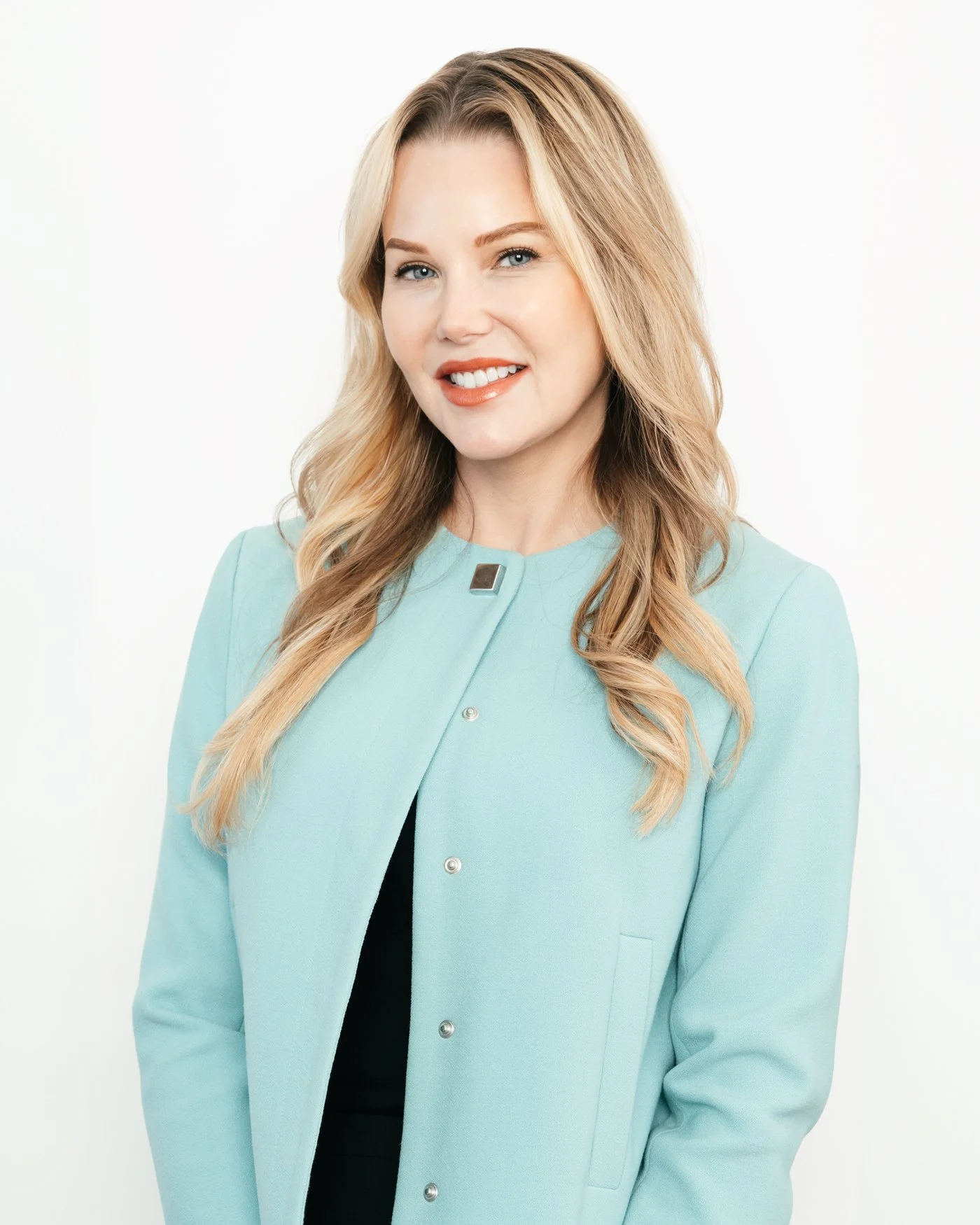 A blonde woman with wavy hair wearing a light blue blazer and an orange lipstick, smiling sweetly against a white background.