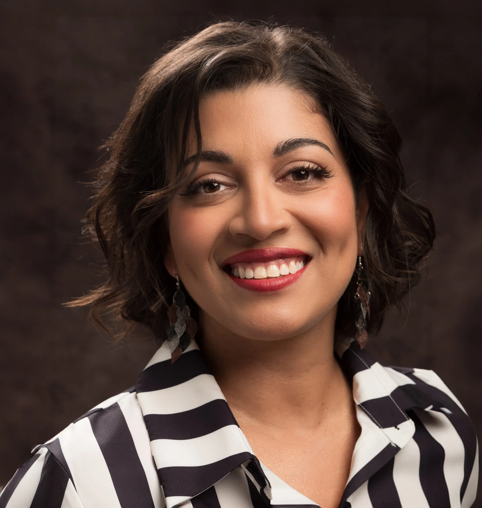 A smiling woman with short, dark hair, wearing a blouse with black and white vertical stripes against a darkbrown, mottled backdrop.