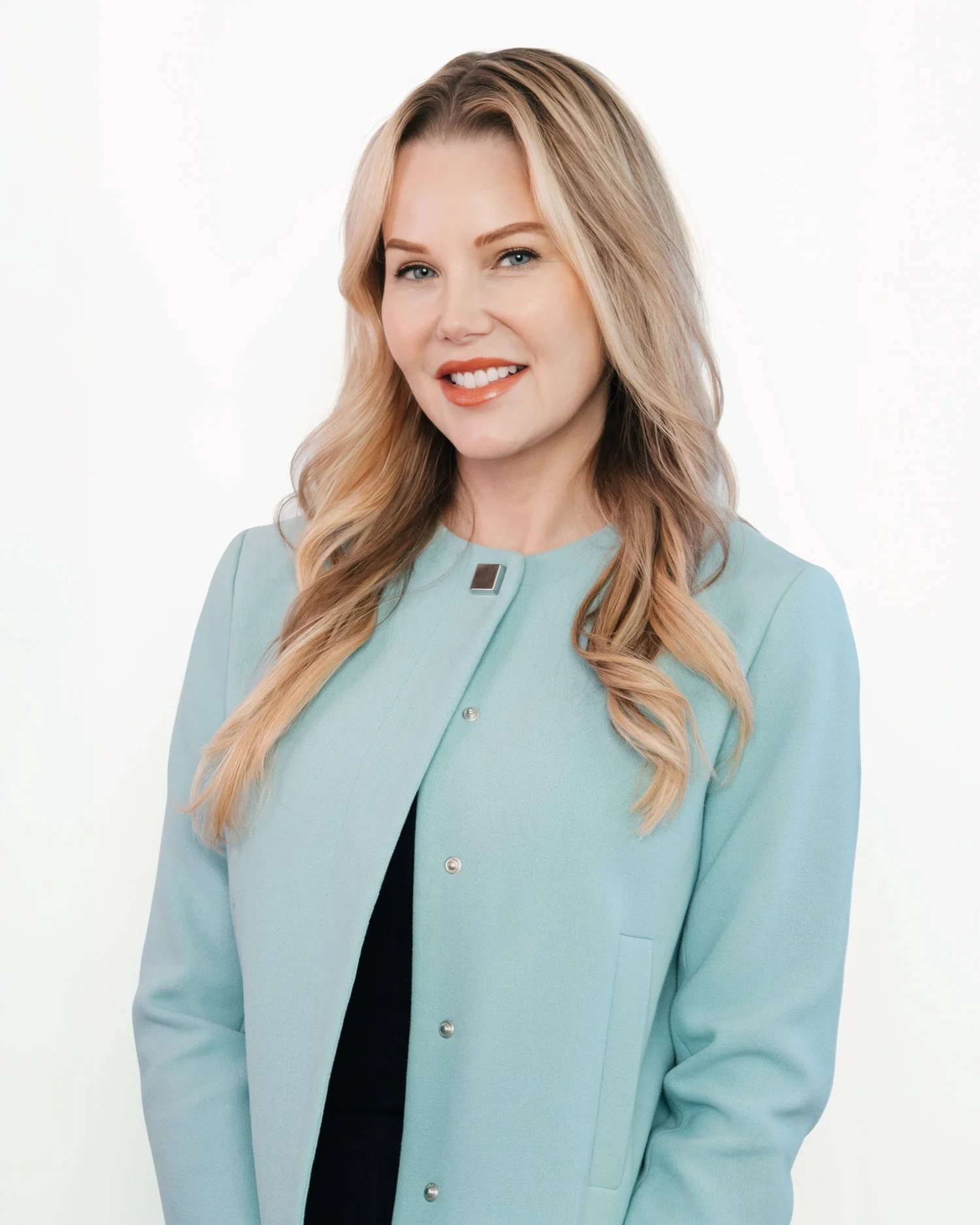 A blonde woman with wavy hair wearing a light blue blazer and an orange lipstick, smiling sweetly against a white background.