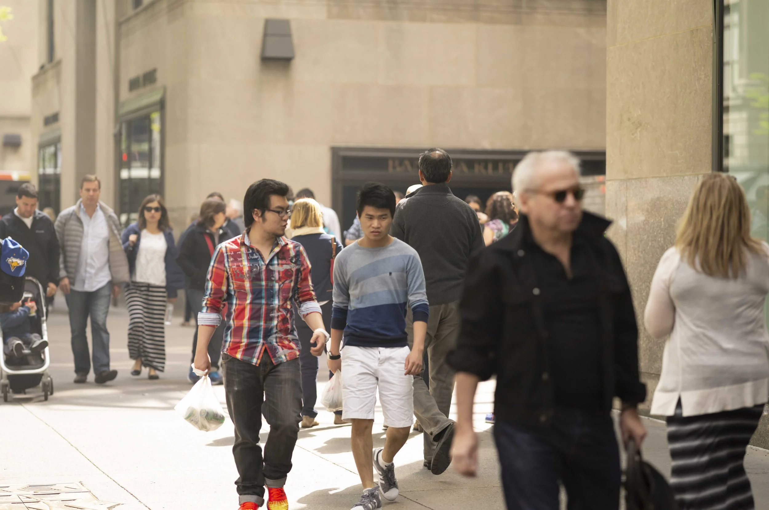 A busy city street with many pedestrians walking in different directions. People are wearing casual clothing, some carrying bags and pushing strollers. The background includes tall buildings with storefronts and windows, indicating an urban area.