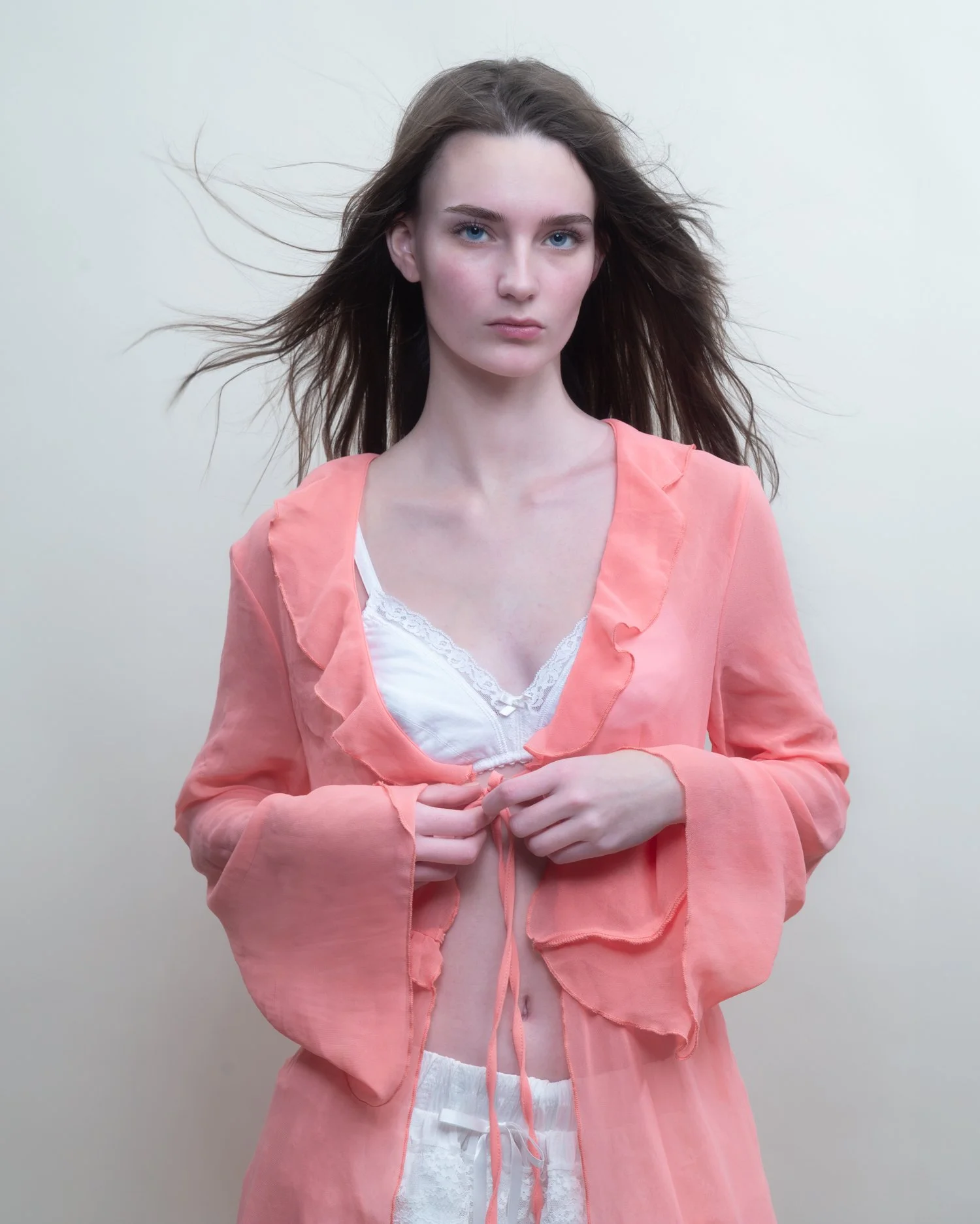 A young woman with long, flowing brown hair, blue eyes, and fair skin, wearing a peach-colored sheer cardigan over a white lace-trimmed top, standing against a plain light background.