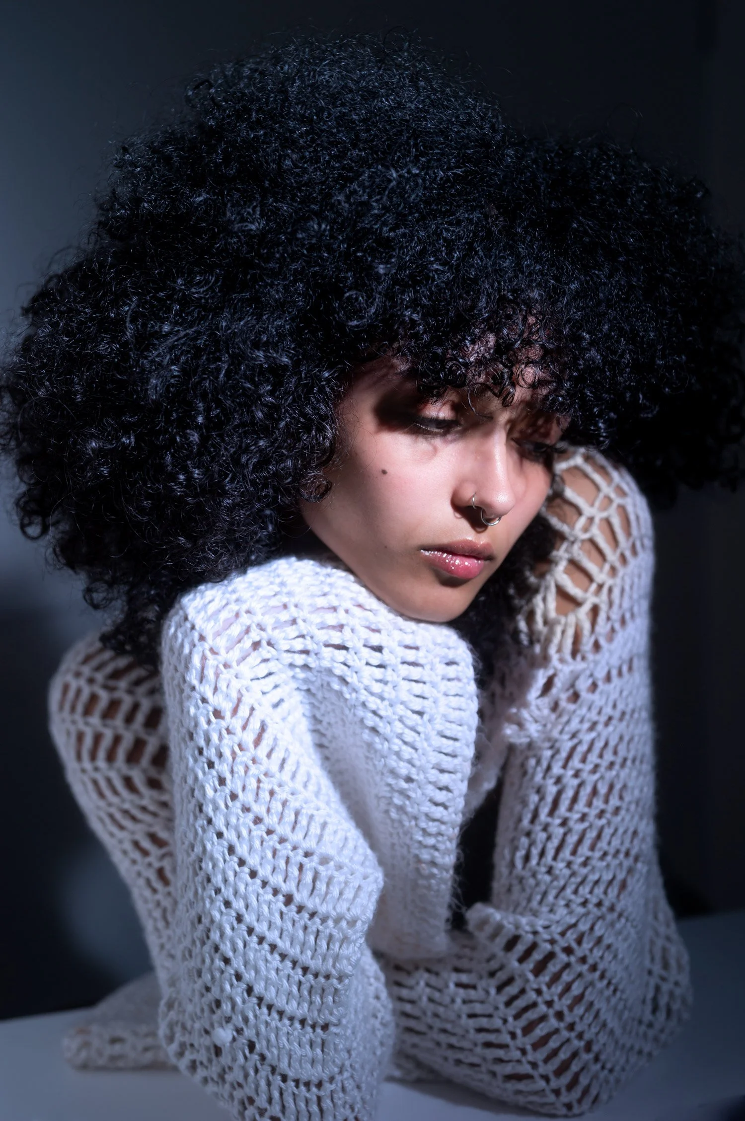 A woman with curly black hair, wearing a white crocheted sweater, resting her head on her arm, looking down with a thoughtful expression.