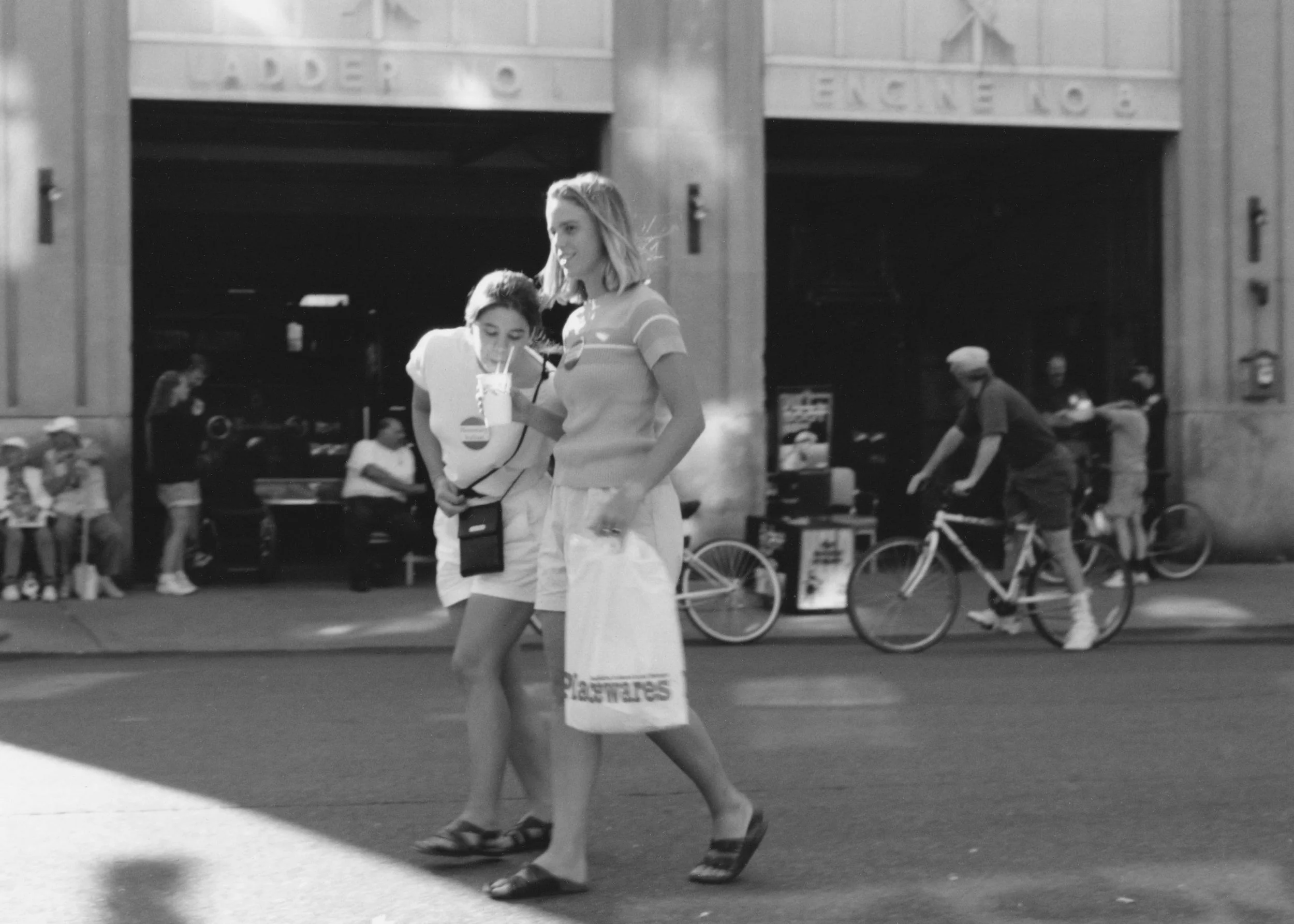 A woman walking on a city street holding a beverage in a cup with a straw. She is wearing a striped t-shirt, skirt, and sandals. In the background, there are people walking, bicycles, and storefronts with signs.