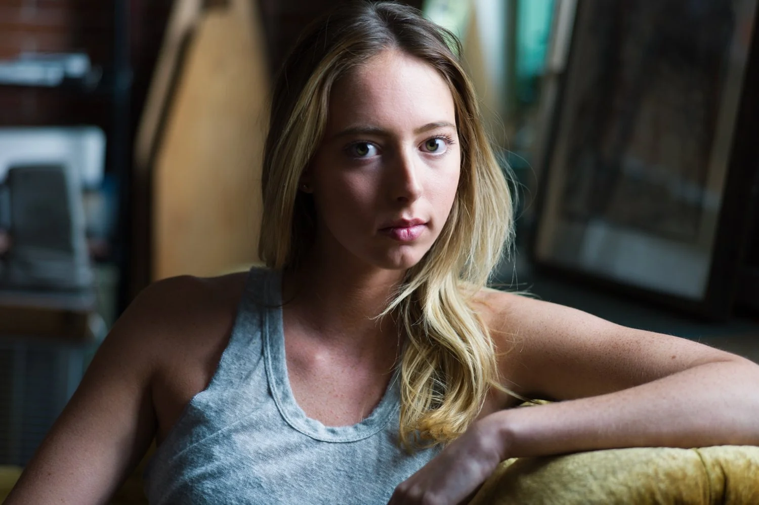 A young woman with blonde hair and green eyes sitting on a yellow chair, looking directly at the camera with a serious expression. She is wearing a gray tank top, and the background features a window and some blurred indoor objects.