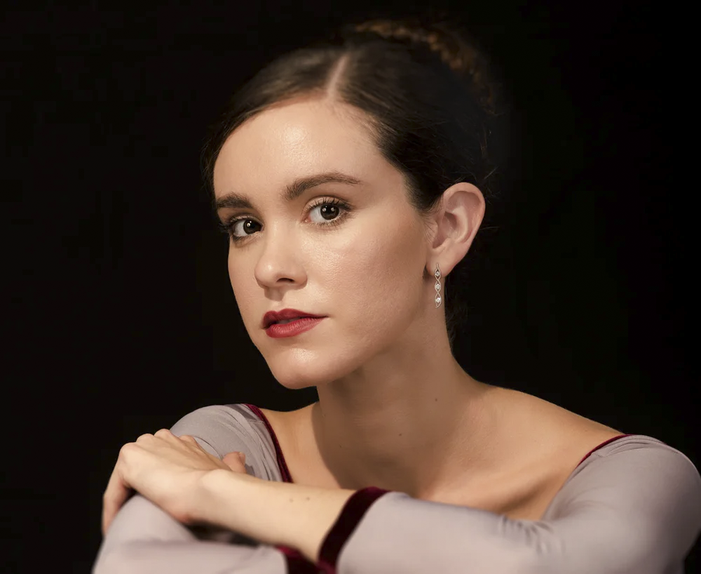 Close-up portrait of a woman with dark hair, fair skin, and red lipstick, wearing earrings and a gray top with maroon accents, against a black background.