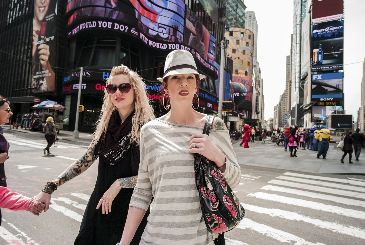 Two women walking across a busy city intersection, holding hands. The woman on the left has long, curly blonde hair, sunglasses, and tattoos on her arm, wearing a black dress. The woman on the right has short, red hair, hoop earrings, a striped shirt