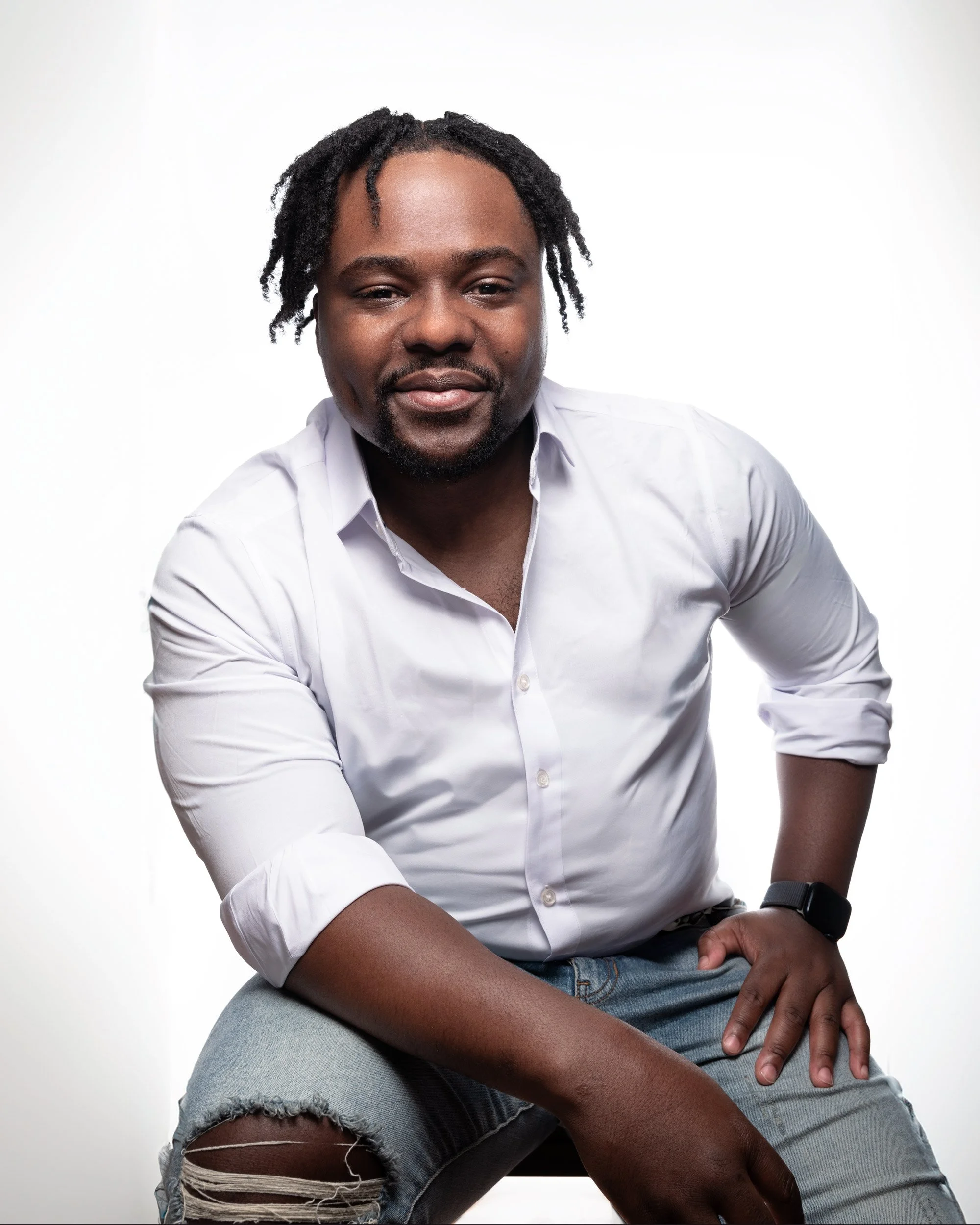 Young Black man with dreadlocks seated on a stool, wearing a white button-down shirt and torn, faded jeans. He looks directly into the camera while resting an elbow across his thigh, while his other hand rests on the opposite leg. The background is s