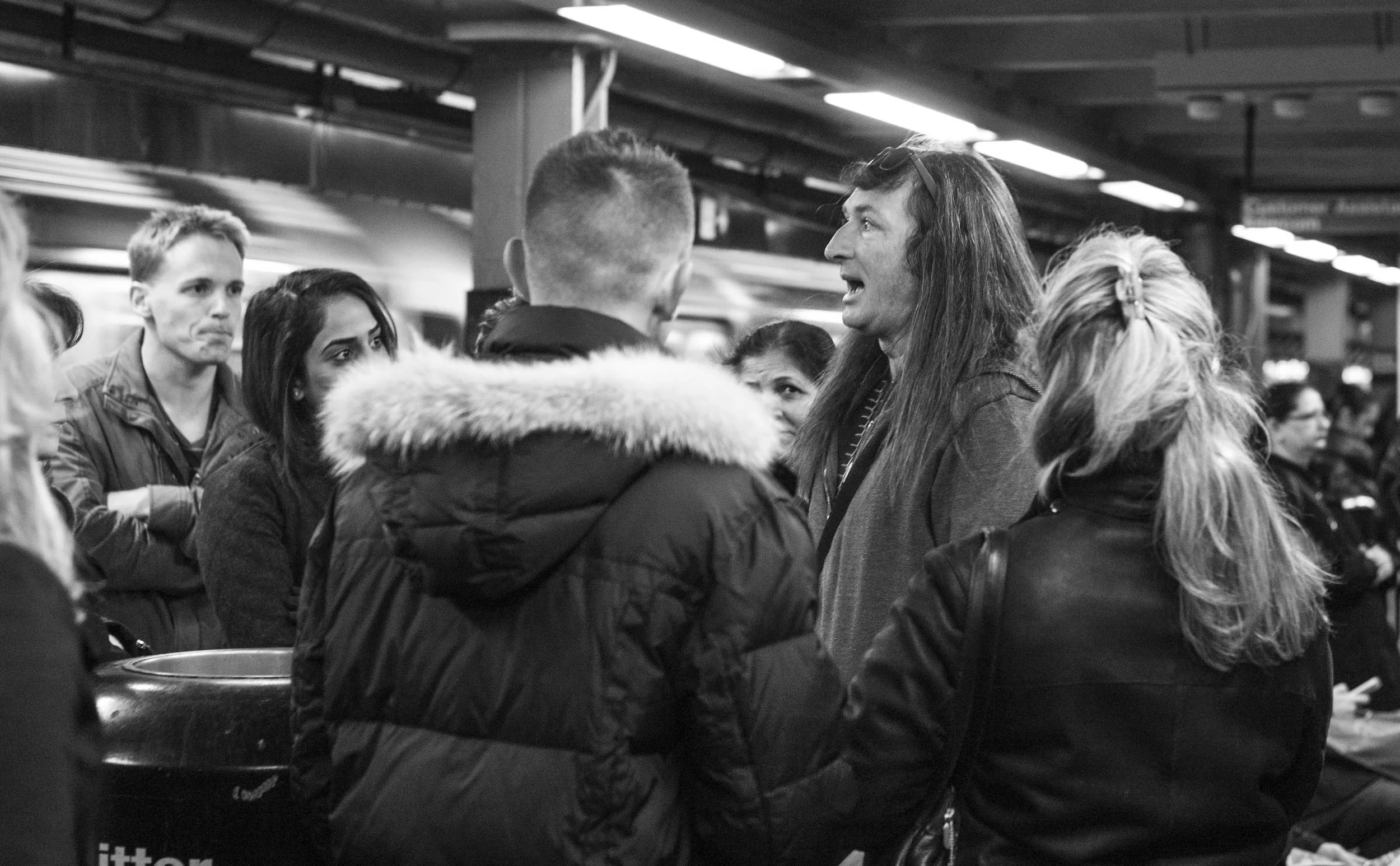 A group of people gathered in an indoor public space, engaging in conversation, with a man with long hair speaking to a man with a short haircut, surrounded by others.