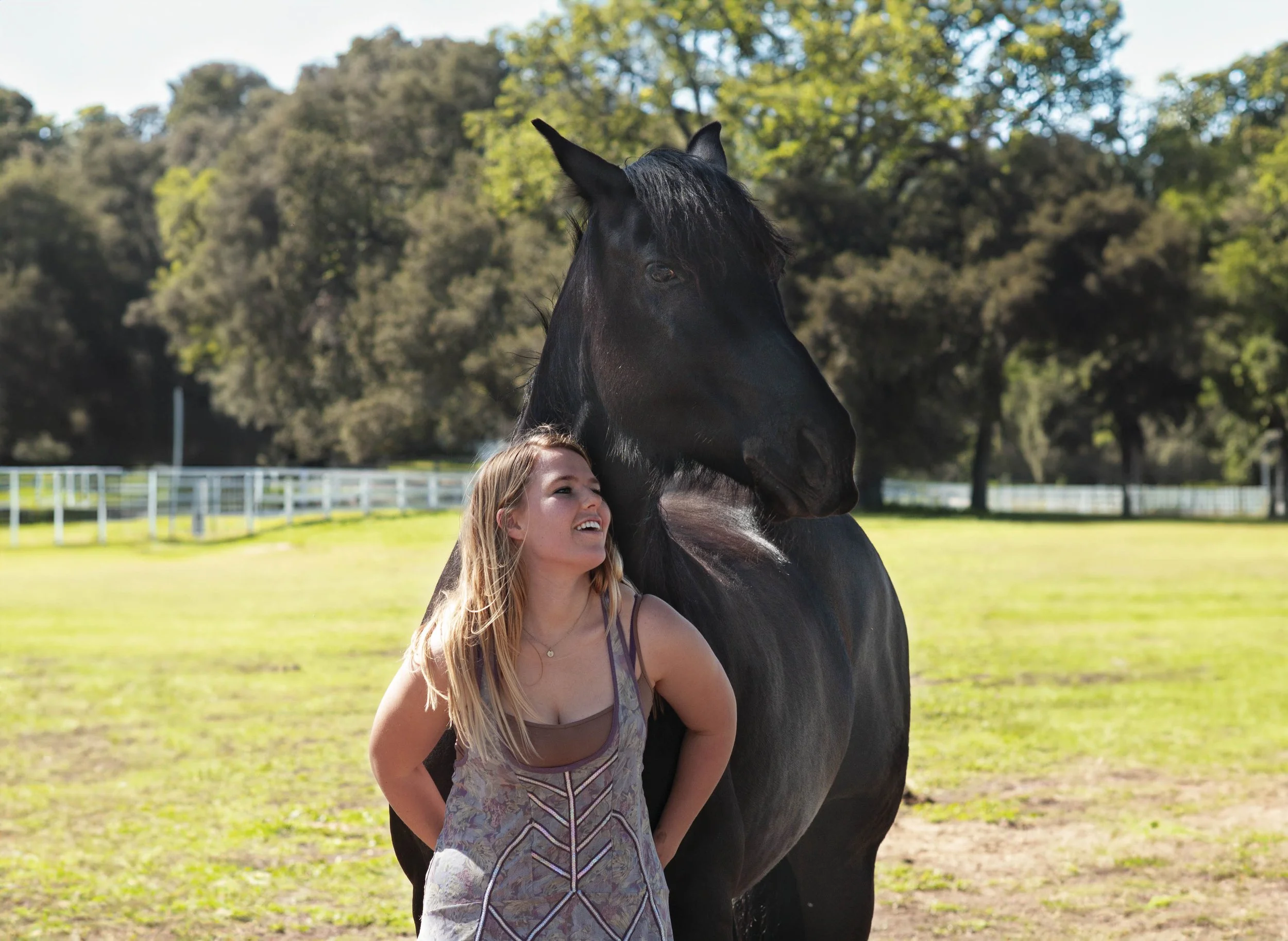 A young woman with long blond hair smiling, standing next to a black horse, outdoors on a sunny day with trees and a white fence in the background.