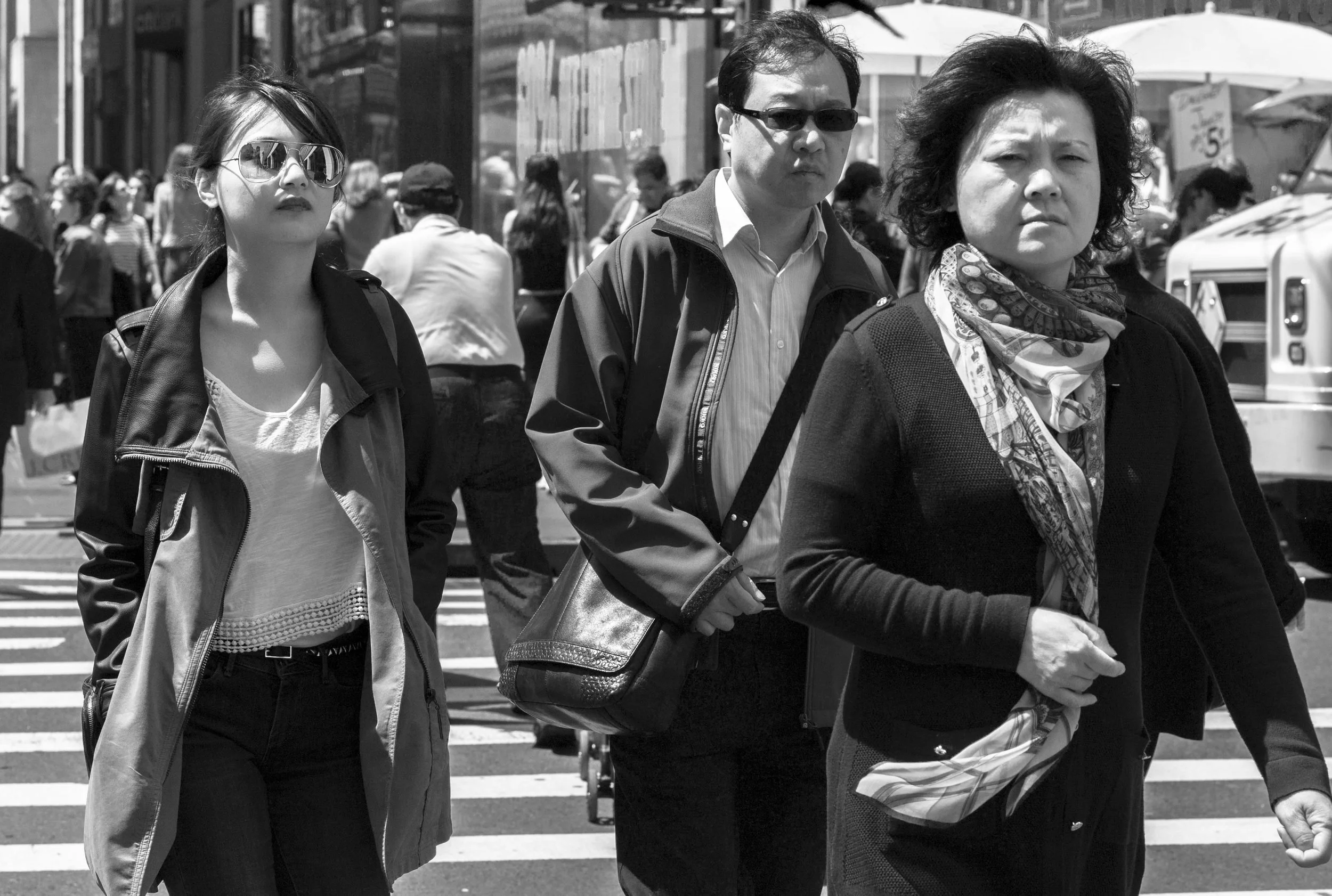 People walking across a street in an urban setting, some wearing sunglasses and carrying bags, in black and white photograph.