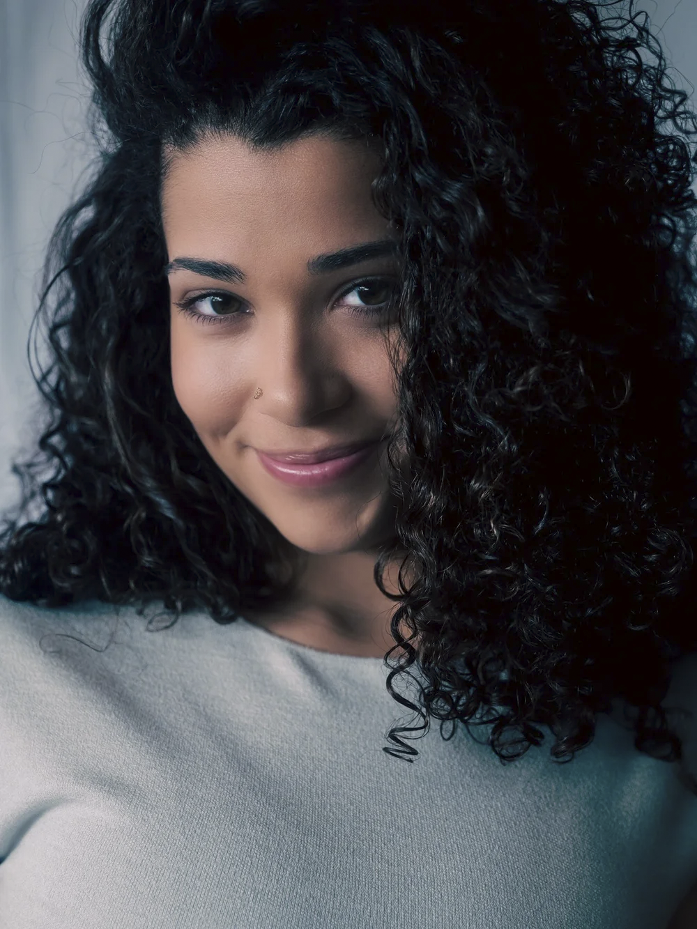 Close-up portrait of a woman with curly black hair, light skin, smile, wearing a light-colored top, and a small nose piercing.